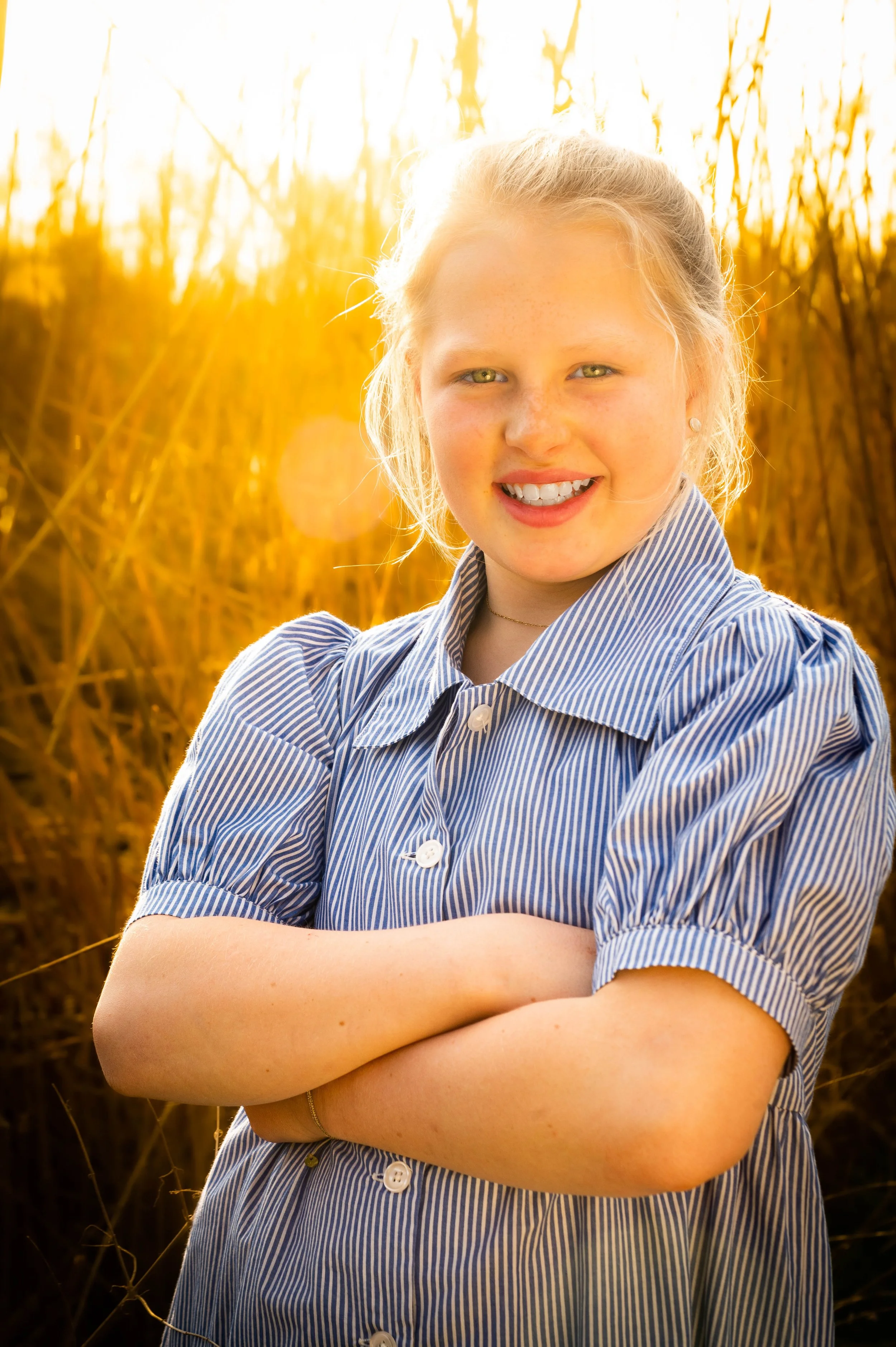 Meisje met blonde haar, gekleed in een blauwe en witte gestreepte blouse, staand met gekruiste armen in een veld met hoge grassen tijdens zonsondergang.