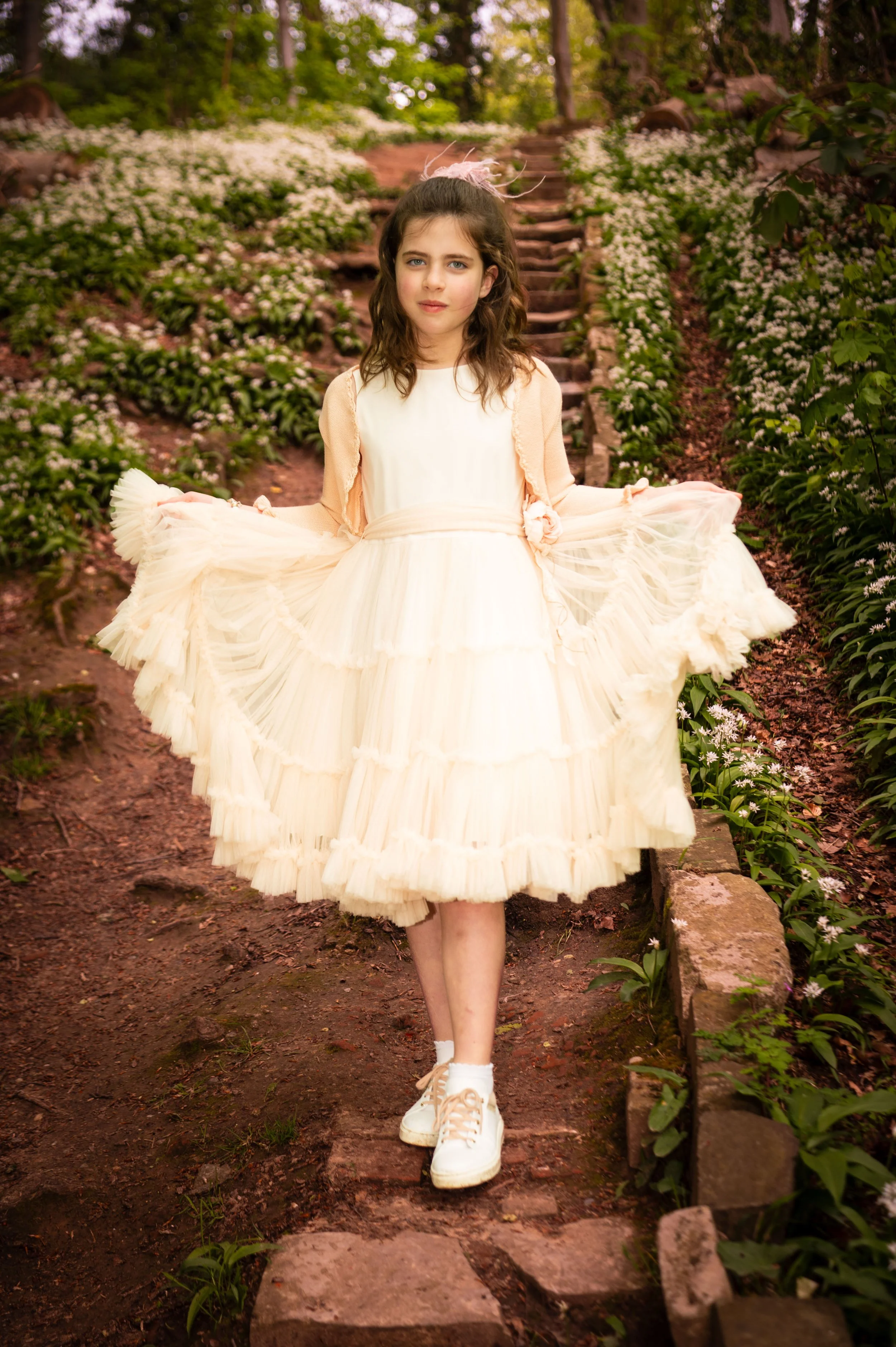 Jonge vrouw in een witte, floersachtige jurk, wandelend op een bosweg met bloemen aan weerszijden.