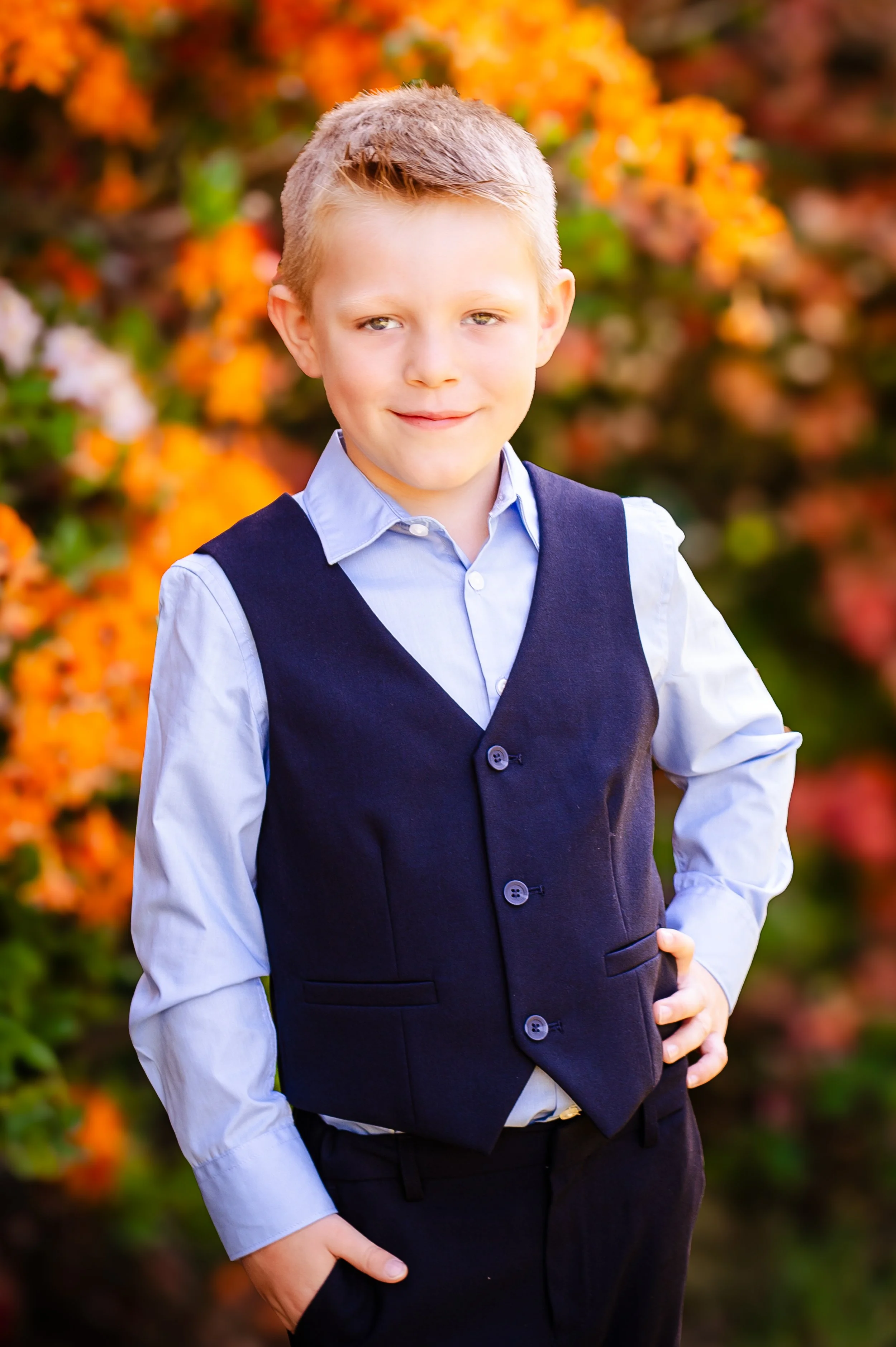 Jongetje met blond haar in nette kleding, poseert voor een kleurrijke bloemenachtergrond.