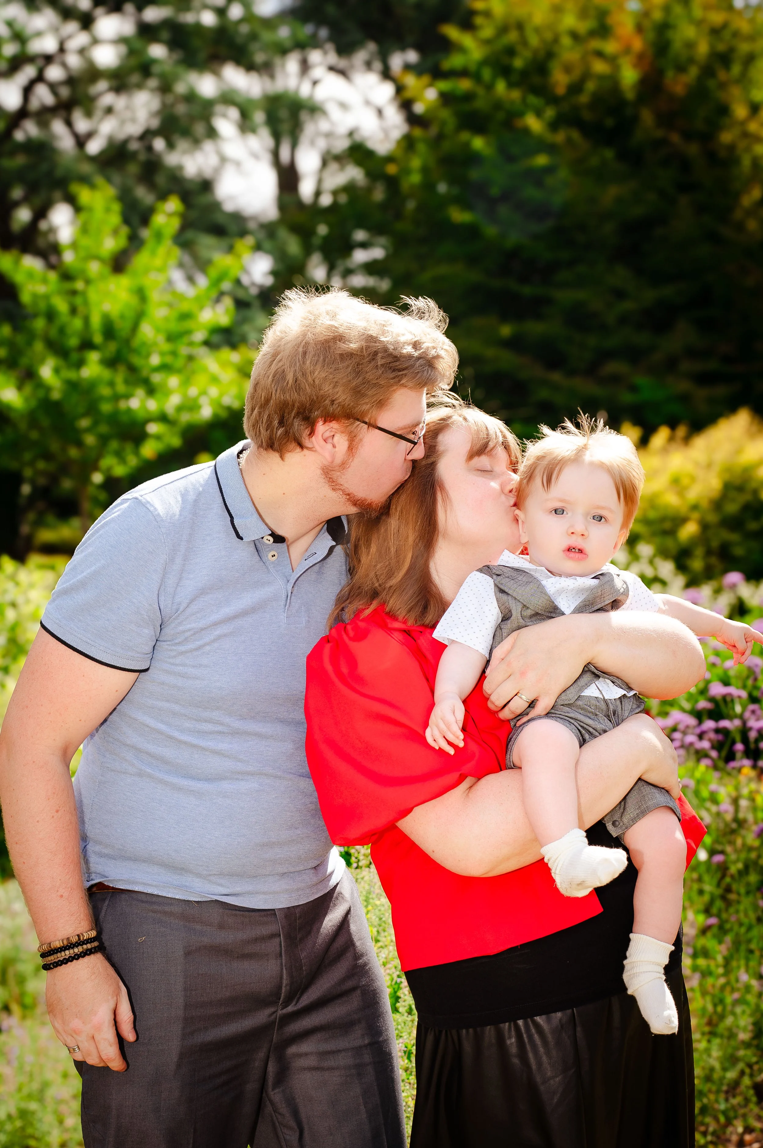 Gezin bestaat uit een man, een vrouw en een baby, buiten in een tuin met groen en bloemen. De man kust de vrouw op het voorhoofd, terwijl de vrouw de baby vasthoudt.