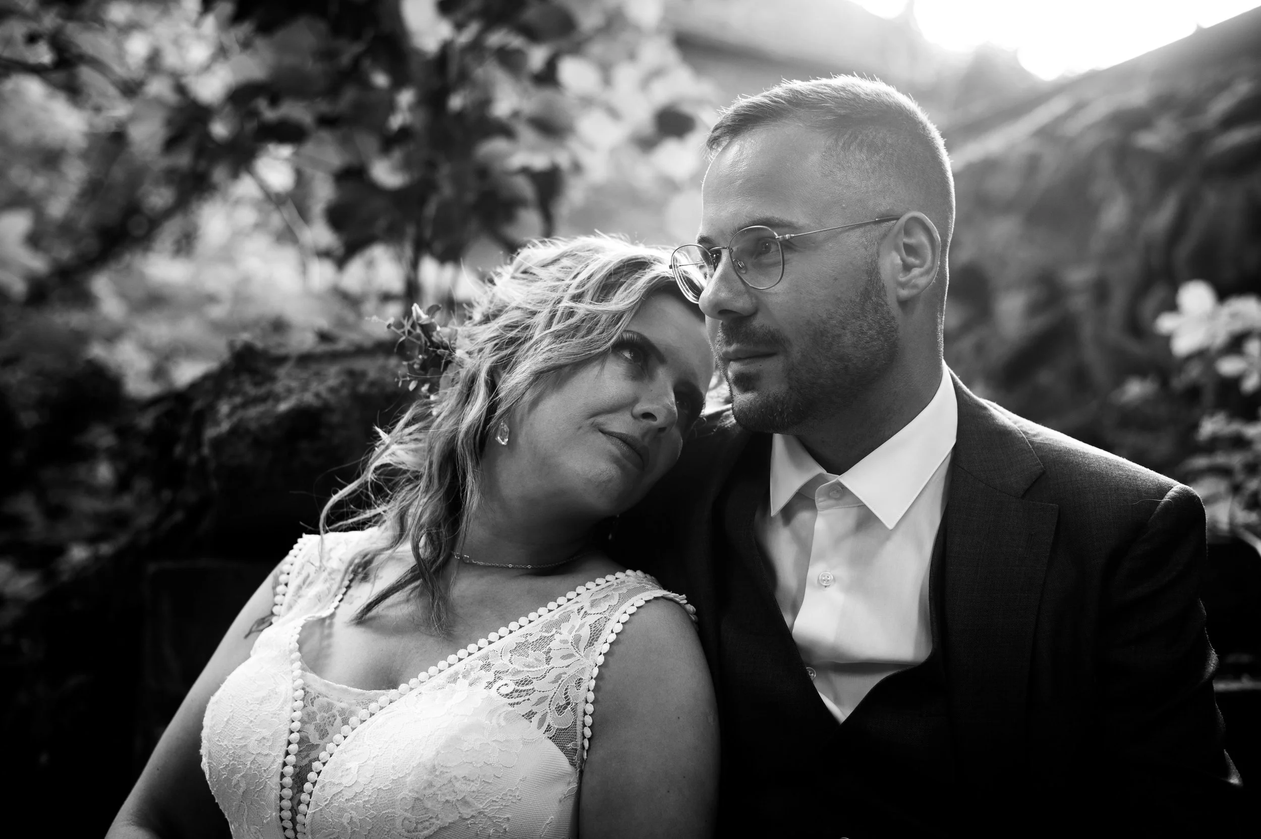 Een zwart-wit foto van een koppel, een vrouw en een man, die intiem naast elkaar zitten in een natuurlijke omgeving met bomen en rotsen. De vrouw kijkt liefdevol naar de man en heeft lang haar, terwijl de man een korte coupe en brillen draagt. Ze lij