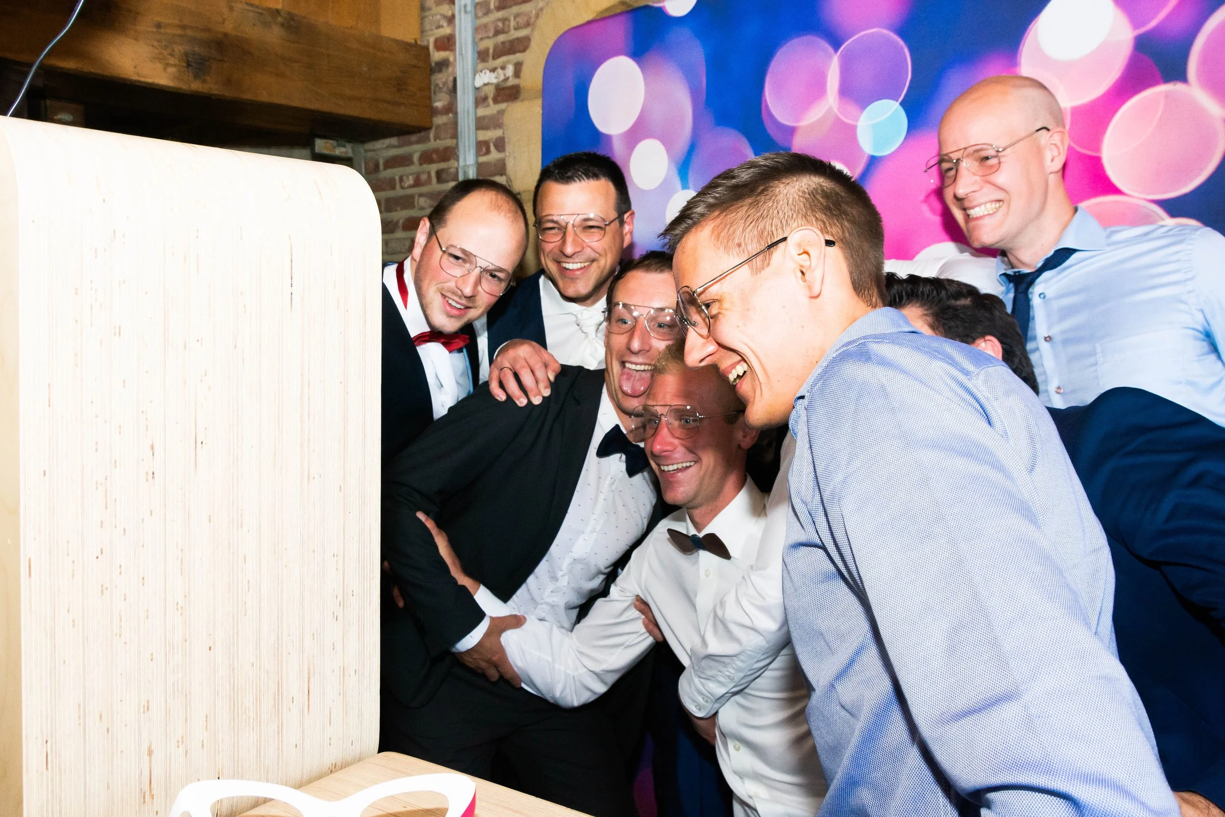 Een groep mannen in formele kleding die samen lachen en poseren voor een foto met een photobooth, in een feestelijke setting.