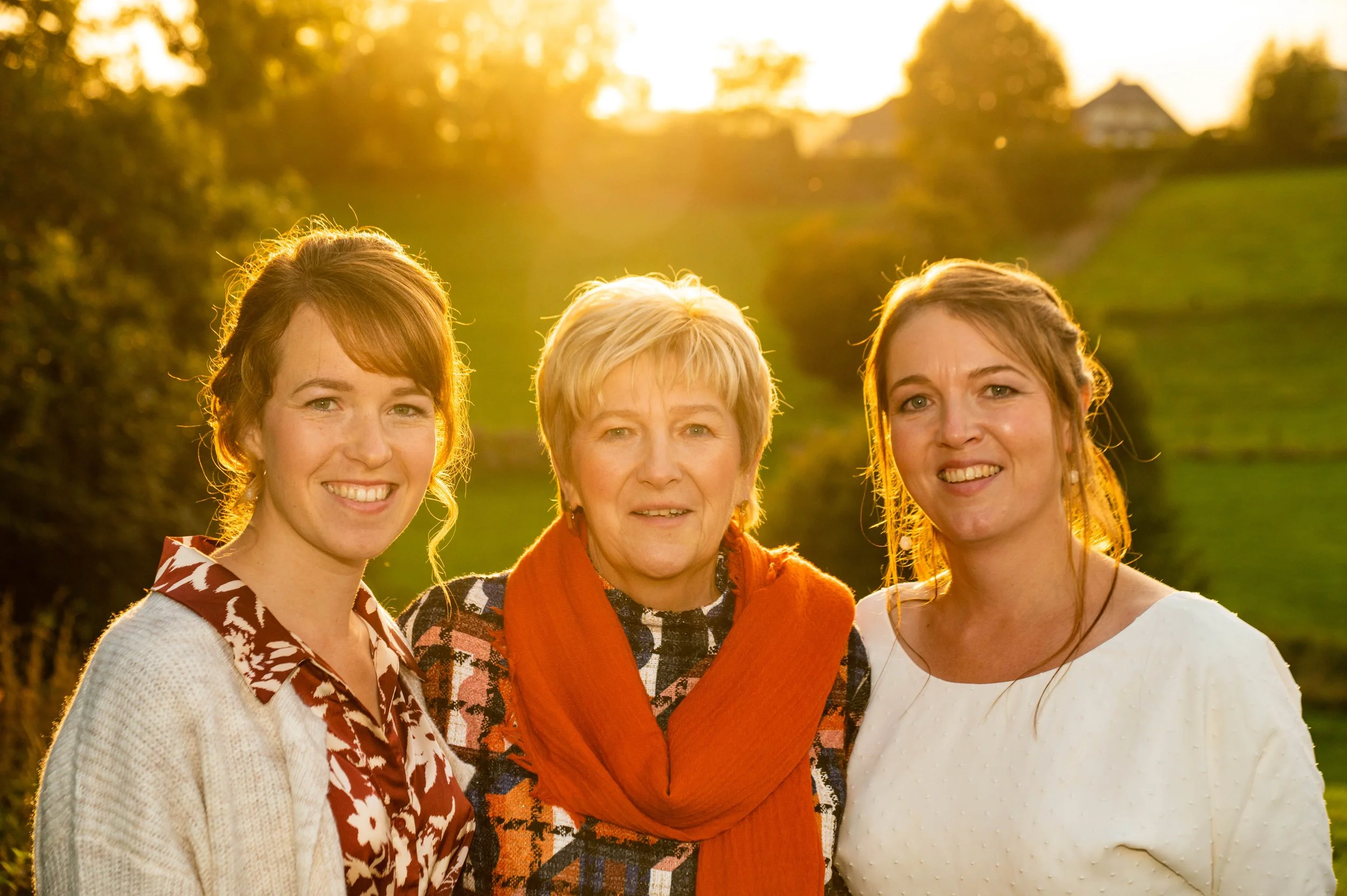 Drie vrouwen lachen samen buiten bij zonsondergang met groene heuvels en bomen op de achtergrond.