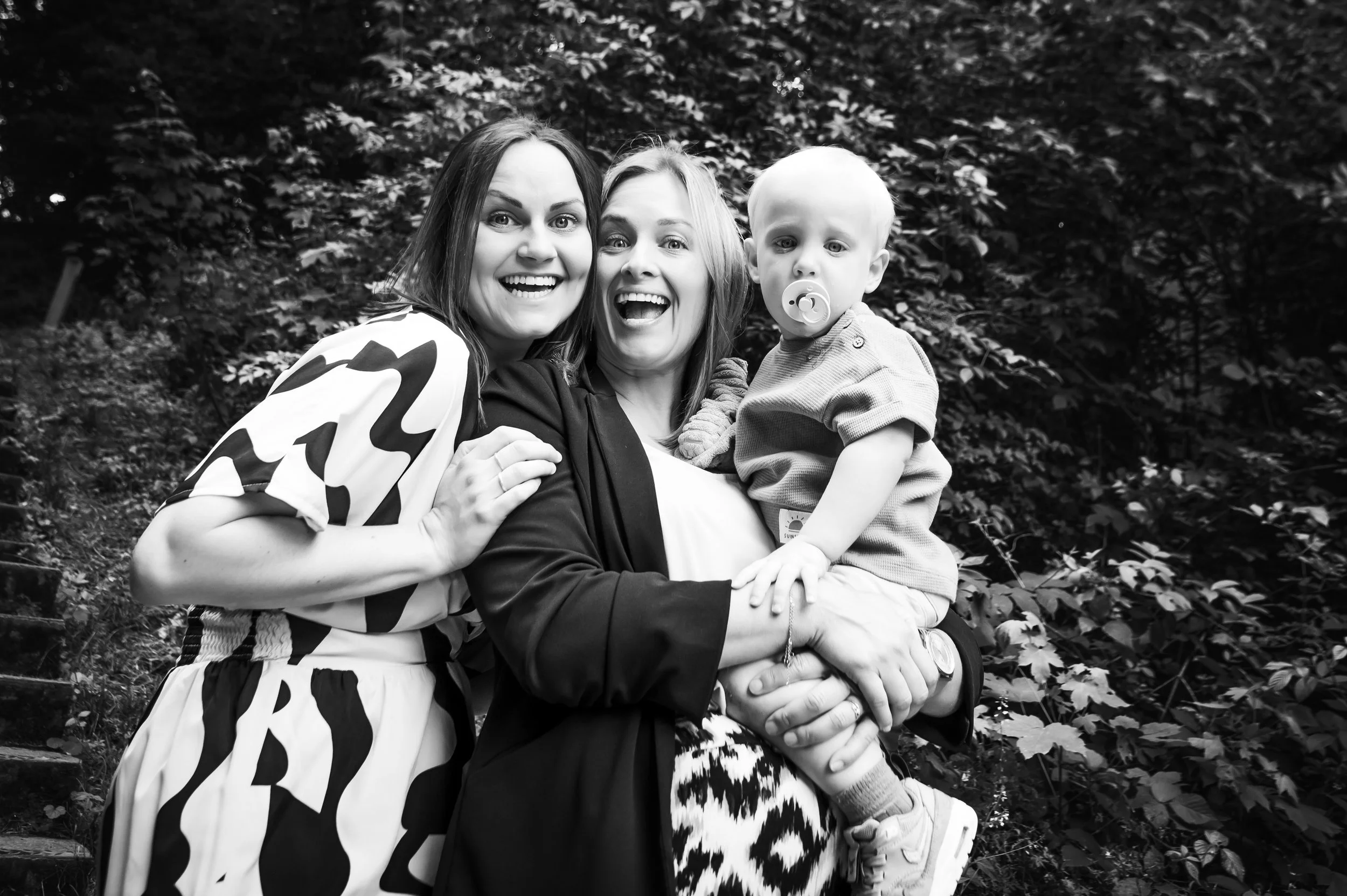 Twee vrouwen en een kind met een pacifier, lachend en knuffelend buiten in een groene omgeving.