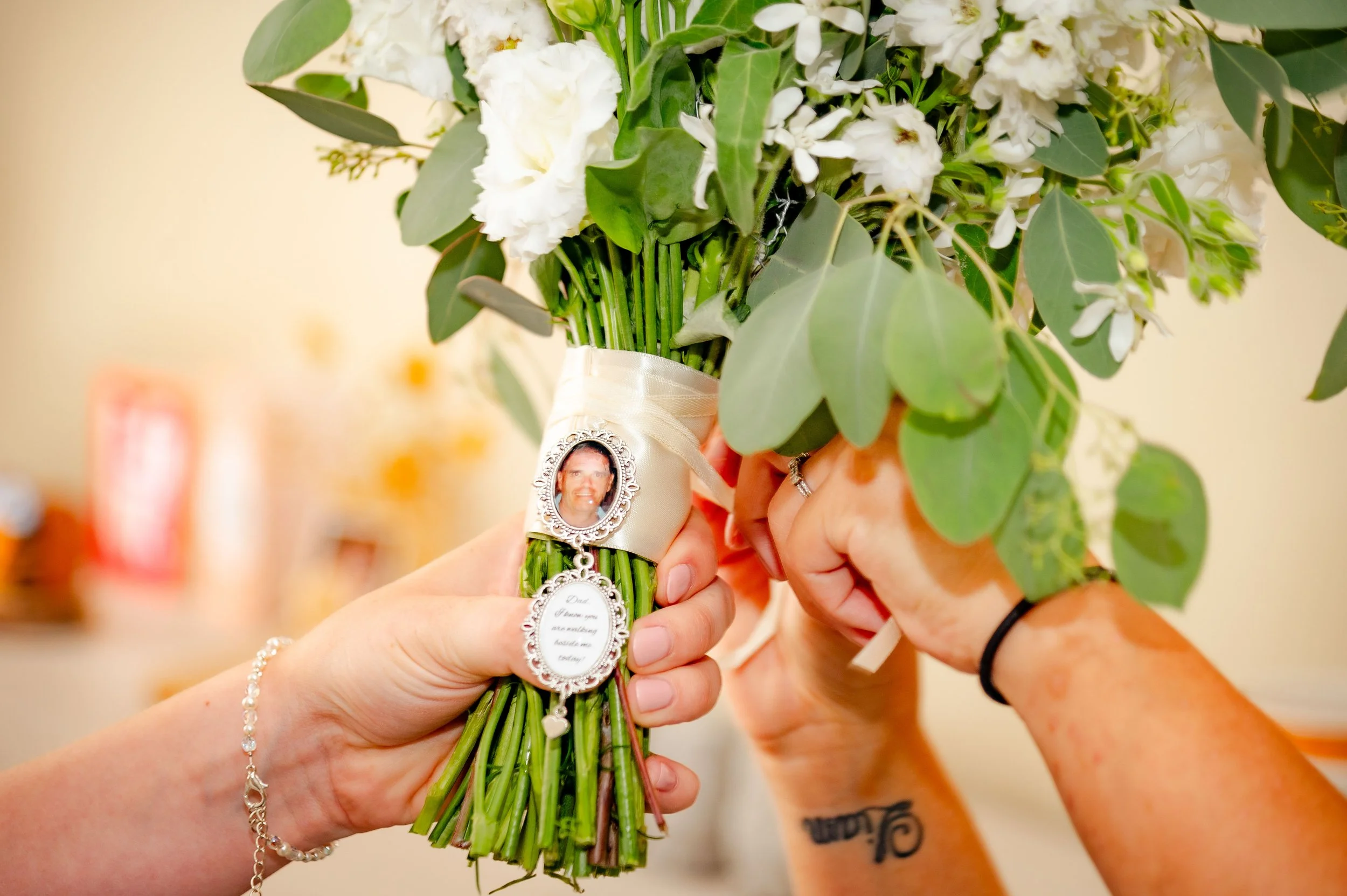 Twee handen houden een boeket witte bloemen vast, versierd met een foto en een bedel. Eén hand heeft een armband en de andere een ring en een zwart polsband.