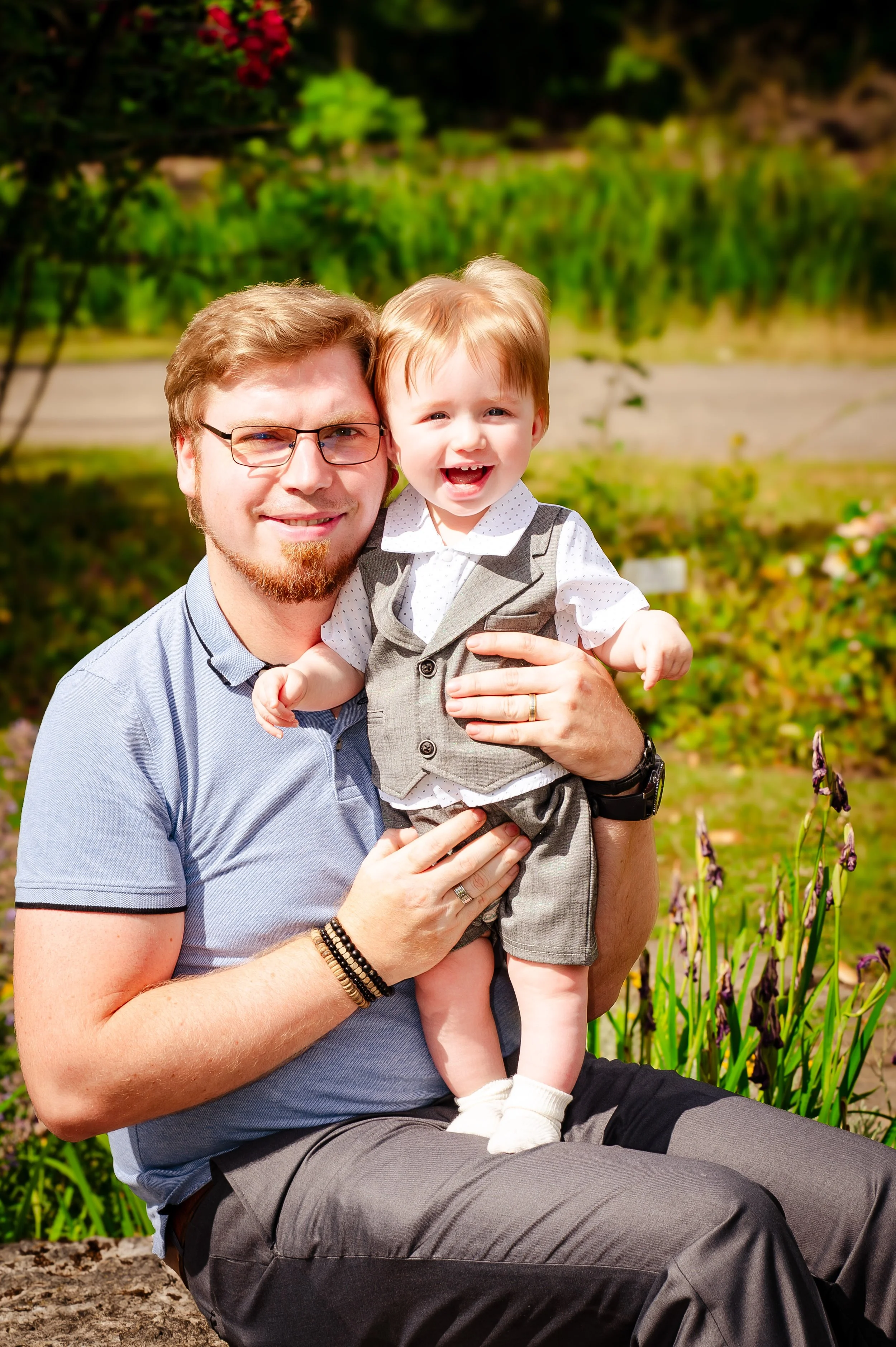 Vader en jonge zoon lachen en poseren buiten in de tuin, omgeven door bloemen en groen.