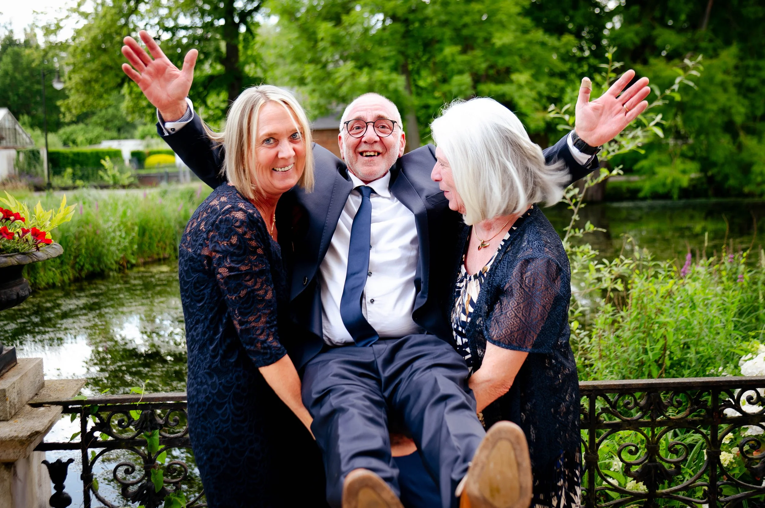 Drie mensen die plezier maken buiten, twee vrouwen en een man, met de man op schoot en de vrouwen aan weerszijden, in een groene tuin met een kleine rivier op de achtergrond.