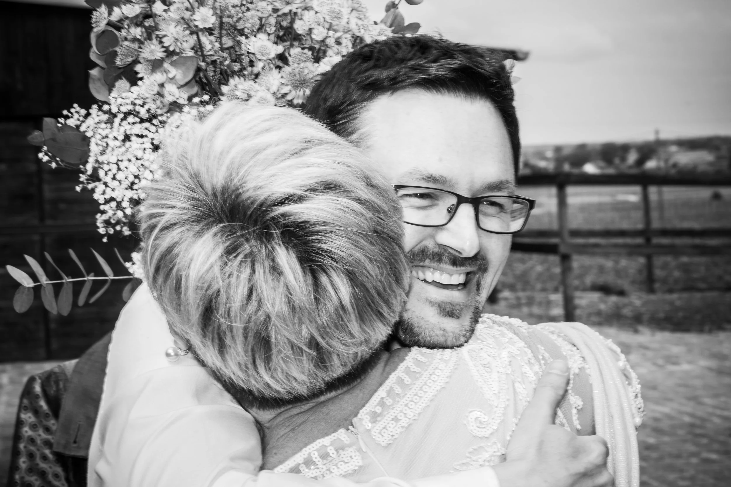 Een man met bruin haar, snor en bril, die glimlacht terwijl hij door een vrouw wordt omarmd. De vrouw heeft licht haar en haar gezicht is niet zichtbaar, omdat haar hoofd in de schouder van de man ligt. Op de achtergrond is een houten hek en een open