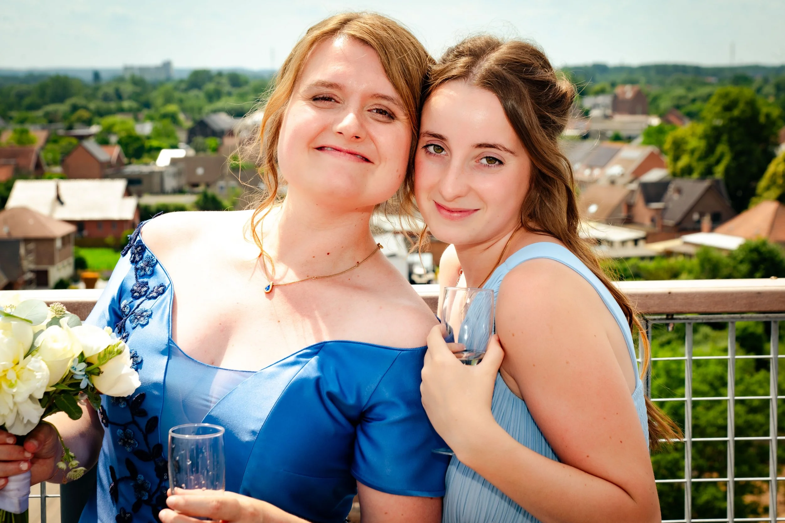 Twee vrouwen op een balkon met een dorp en groene omgeving op de achtergrond, waarbij ze proosten met wijn en glimlachen.