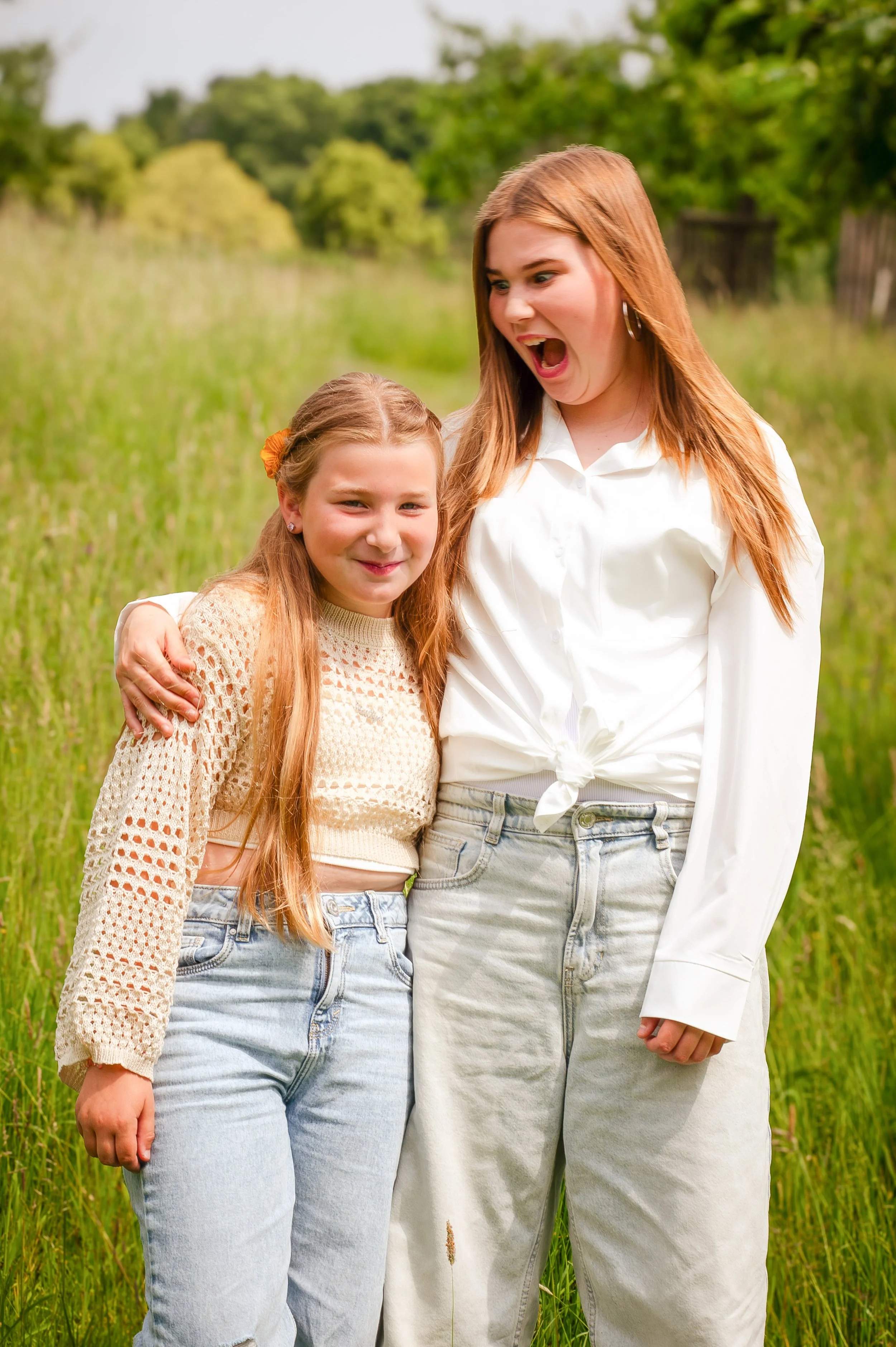 Twee jonge meisjes in een groen veld, één met lang blond haar en een gebreide trui, de andere met lang kastanjebruin haar en een witte blouse, die samen lachen en poseren voor de camera.