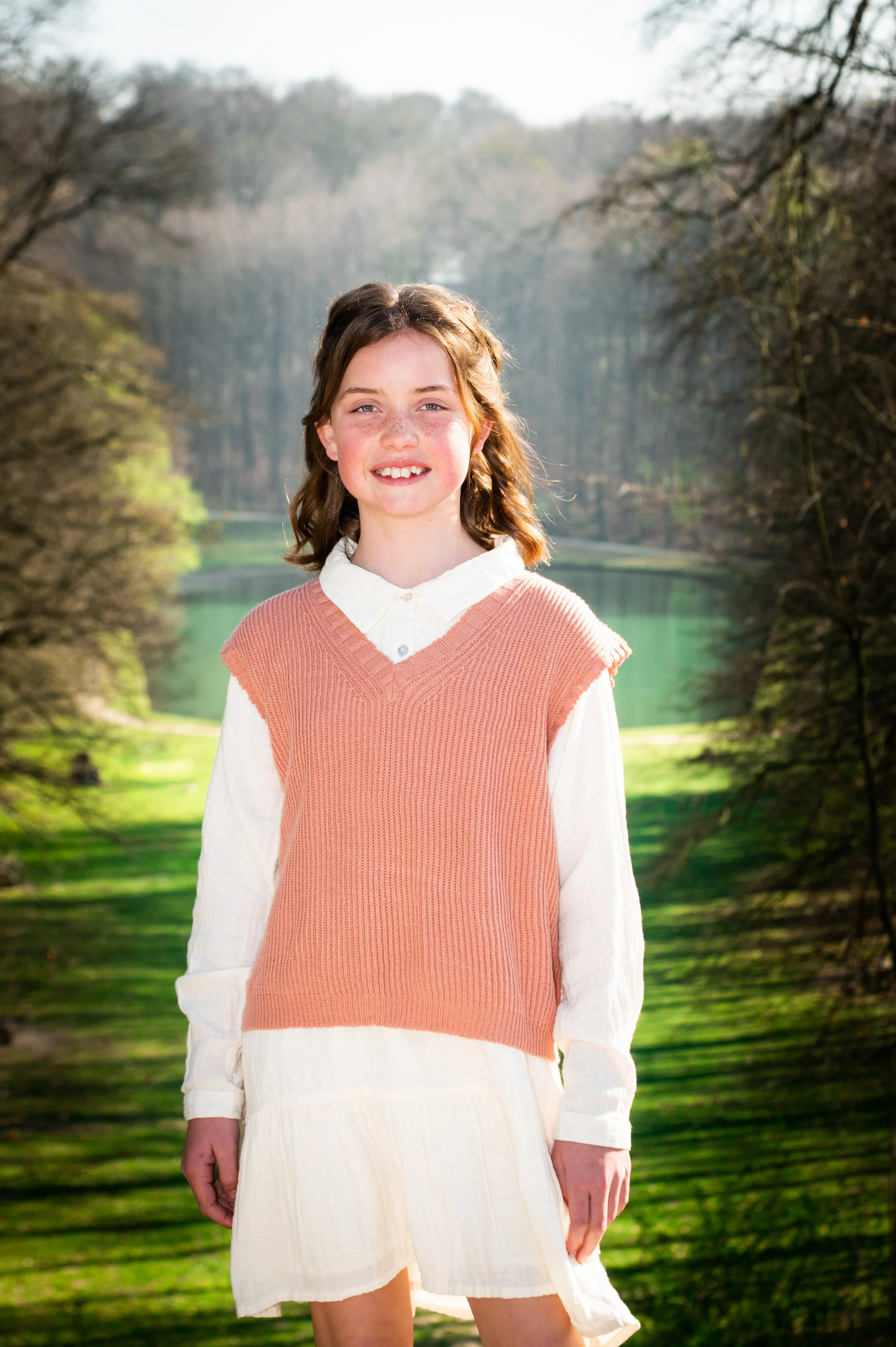 Een glimlachende jonge vrouw in een lichte jurk en een oranje gebreide top, staande voor een achtergrond van een rivier en bomen in het voorjaar.
