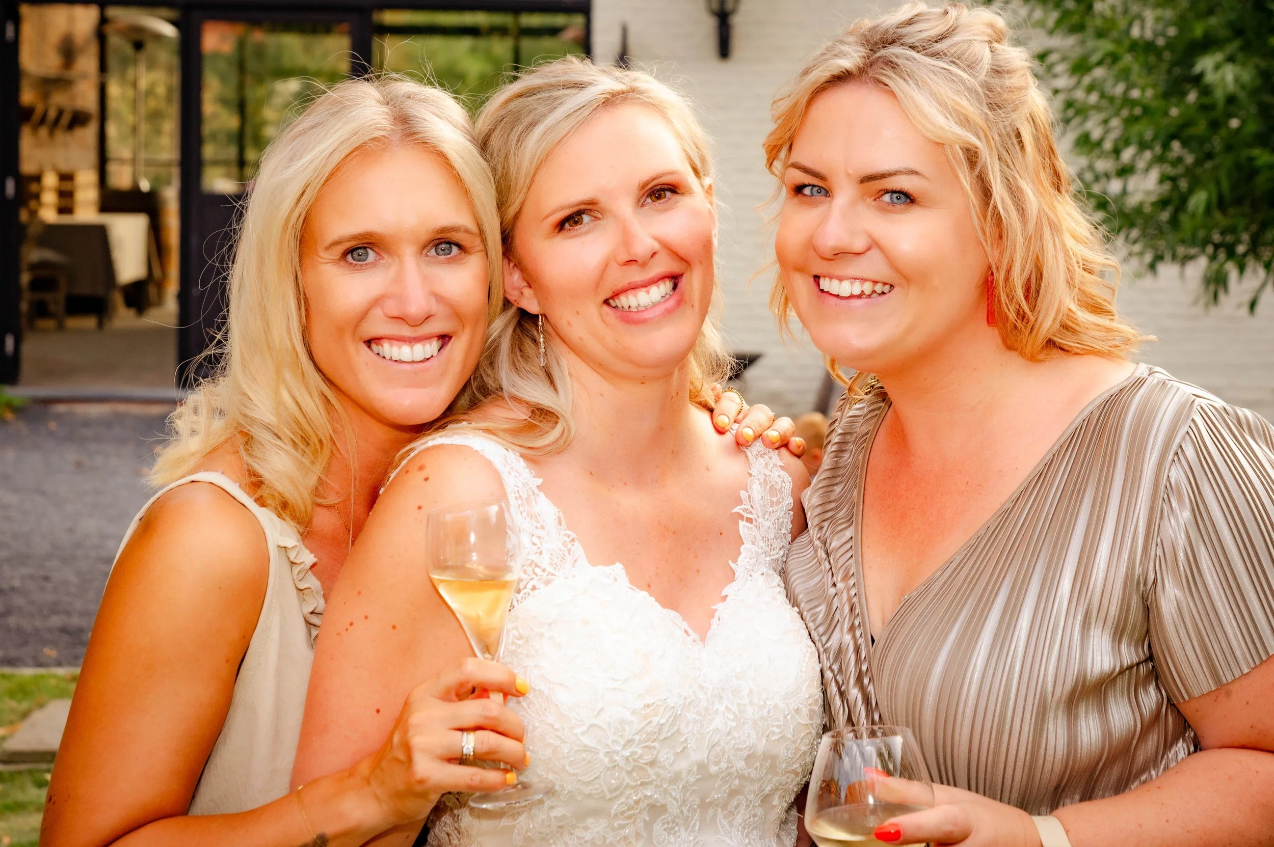 Drie vrouwen glimlachen en drinken wijn op een feest, één vrouw draagt een witte jurk.
