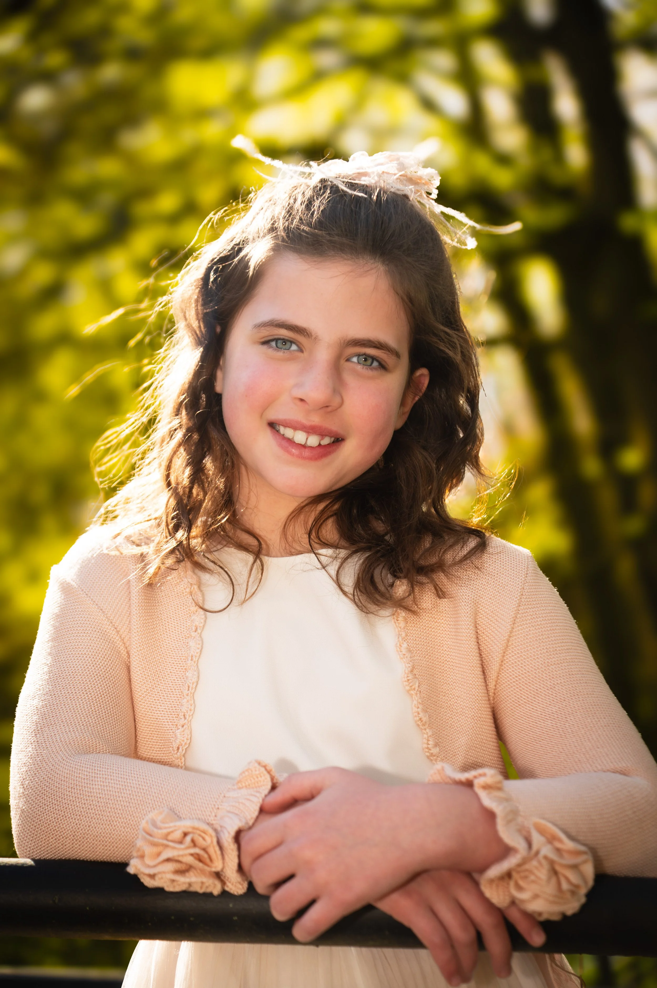 Portret van een lachende jonge vrouw met blauwe ogen en bruin, golvend haar, die in een park staat in de zon, met een achtergrond van groene bladeren.