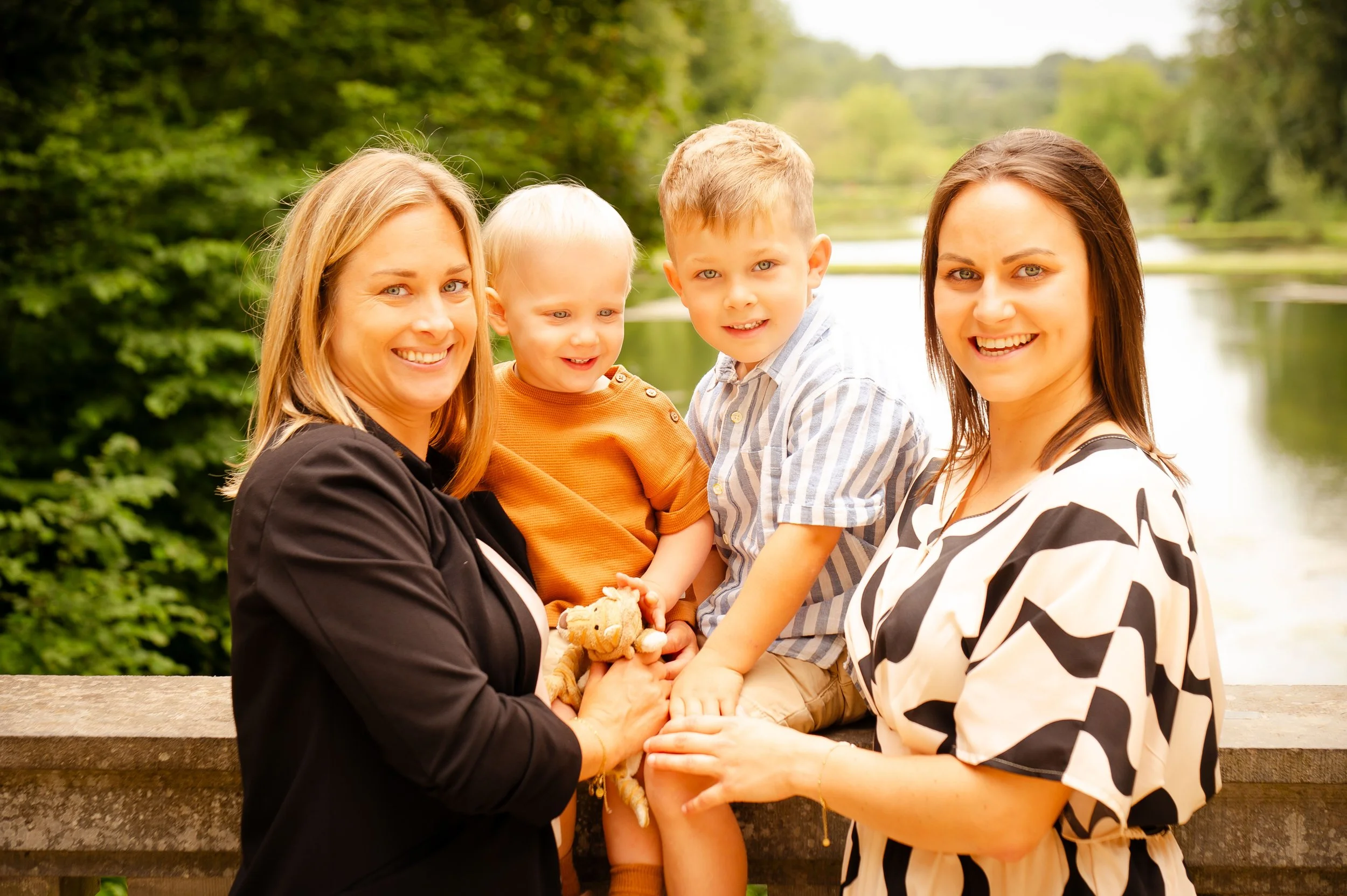Vrouw met blond haar en een zwarte jas met twee jonge jongens en een jonge vrouw, staand op een houten brug bij een meer met groenbos op de achtergrond, smiling aan de camera.