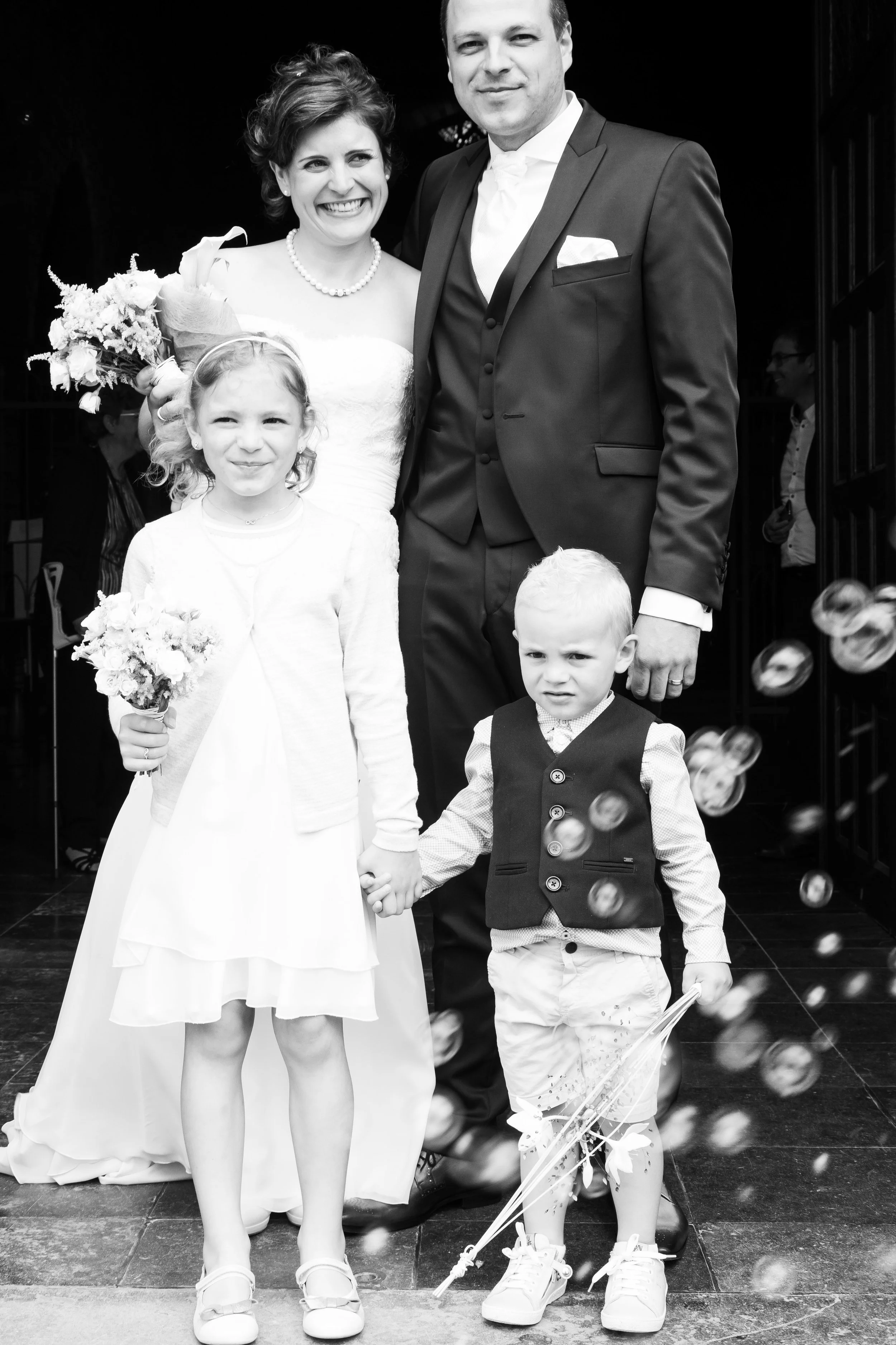 Een zwart-wit foto van een bruid en een bruidegom samen met twee jonge kinderen op een huwelijksfeest. De kinderen houden elkaars hand en de bruid houdt een bloemenboeket.