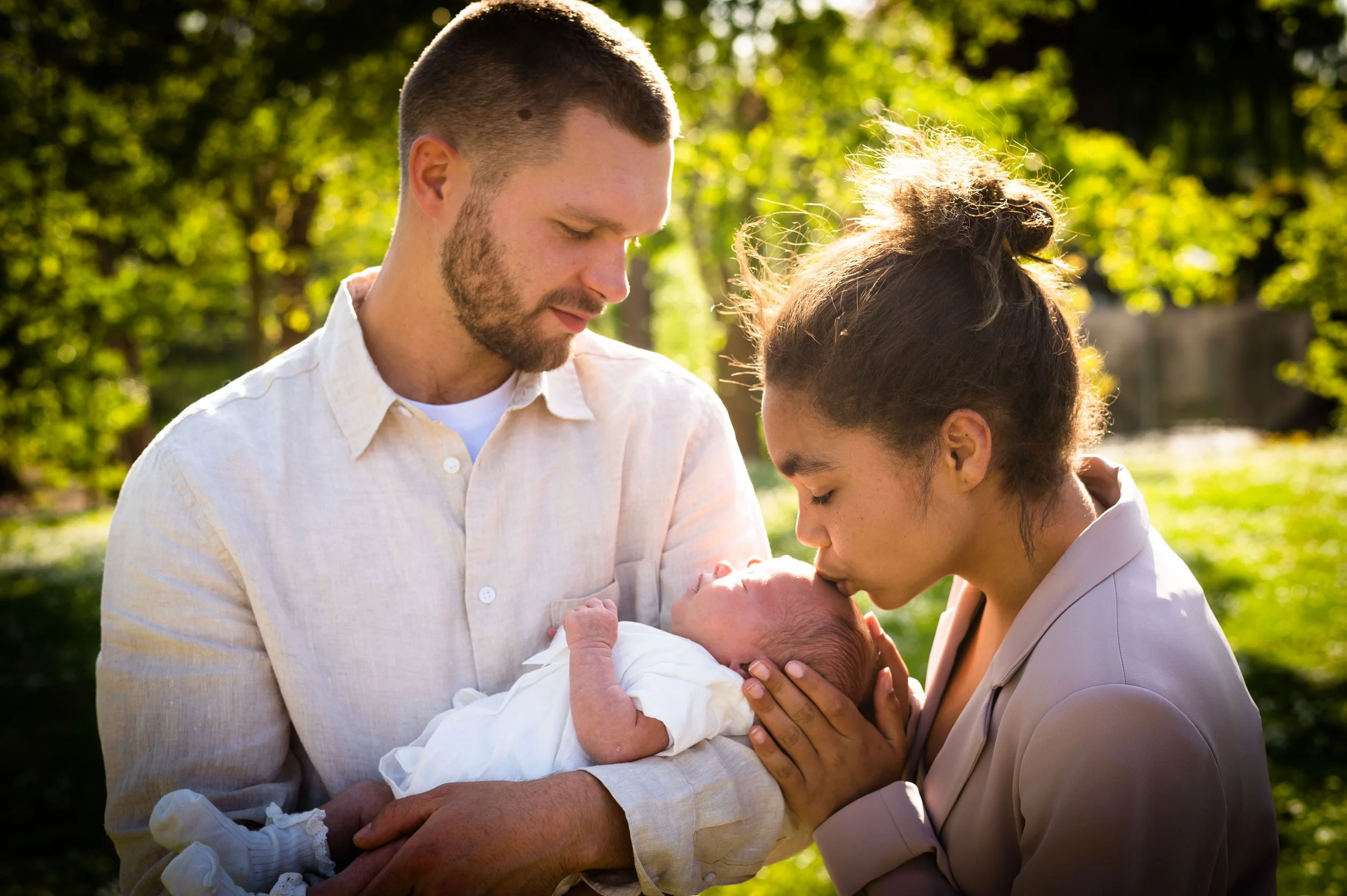 Een jonge man en vrouw in een park met een baby, met de man die de baby vasthoudt en de vrouw die de baby kust, omgeven door groene bomen en zonlicht.