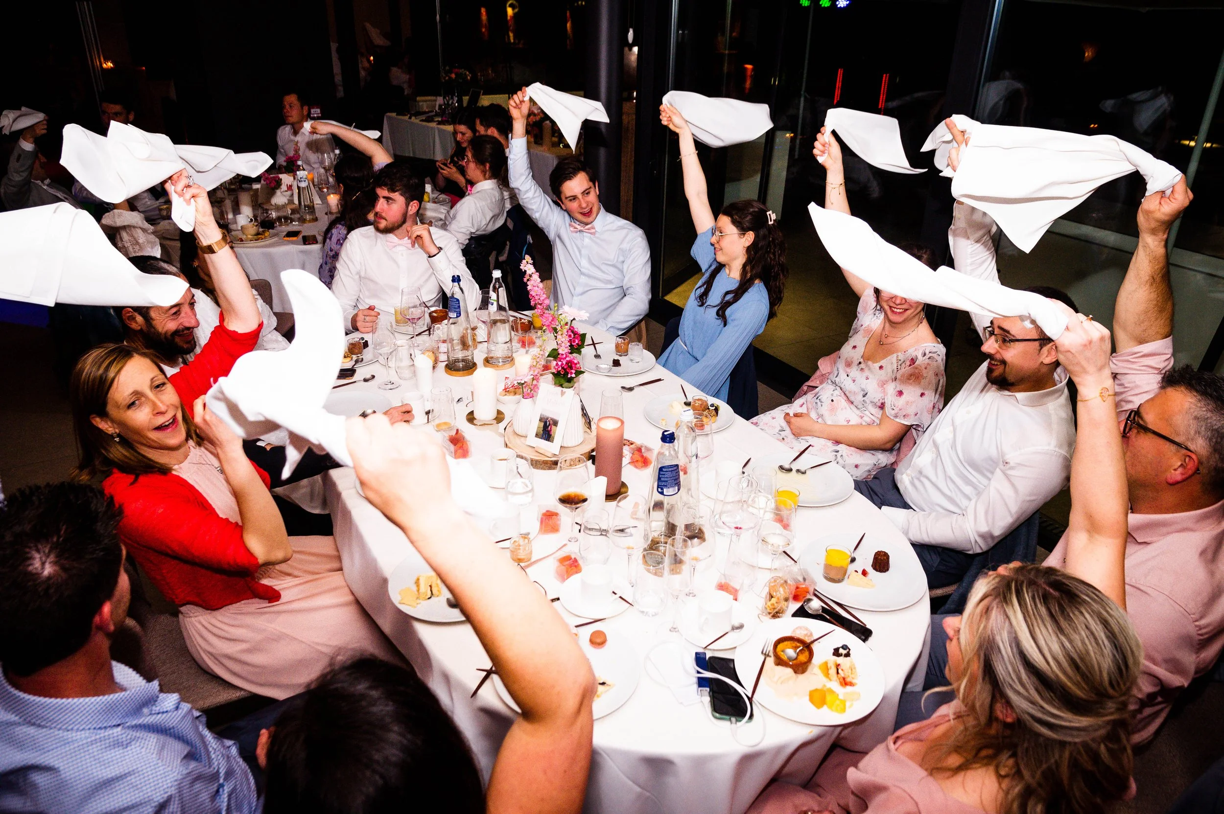 Gasten aan een feestelijke tafel met witte tafellakens, die lachend en plezierig interactie hebben, terwijl ze witte servetten in de lucht zwaaien.