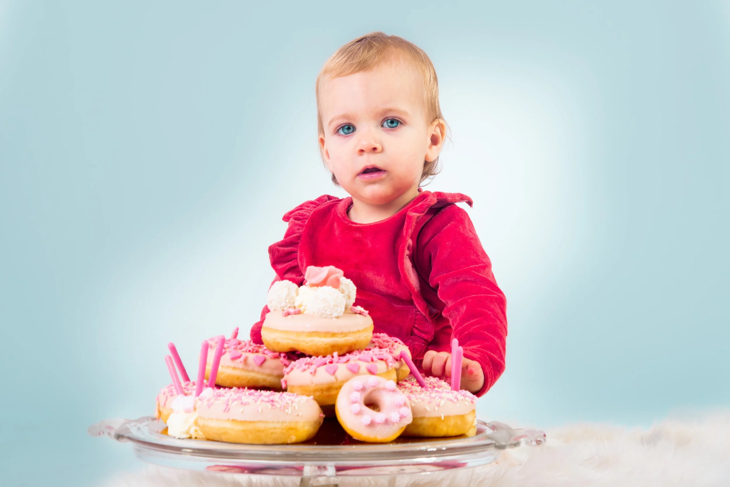 Kleine jongen met blauwe ogen en blond haar zit achter een tafel met een verjaardagskaart die aan een vork hangt, omringd door diverse roze en witte donuts met decoraties.
