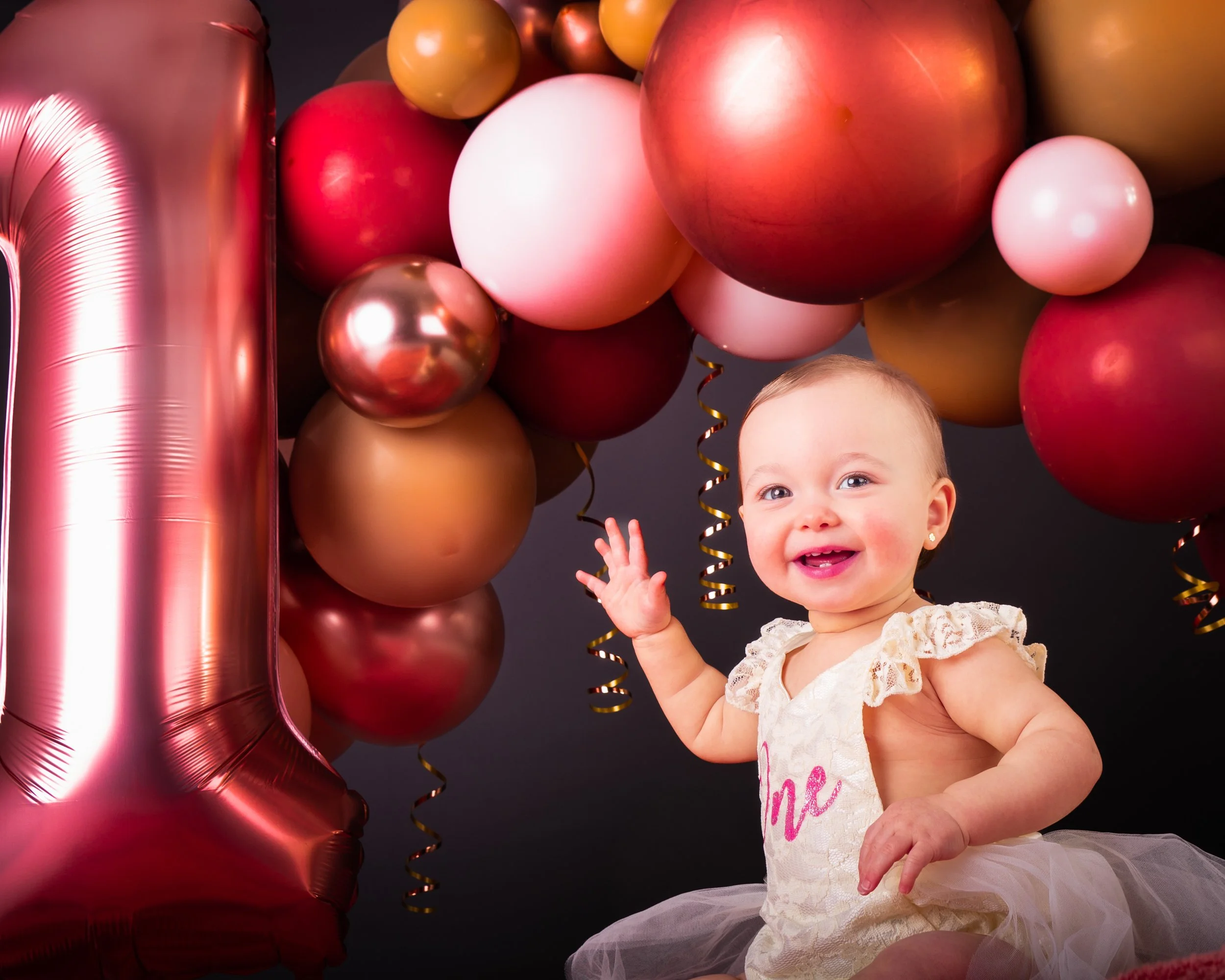 Een lachende baby op haar eerste verjaardag met ballonnen in roze, goud en koper, en een grote roze nummerballon '1'.