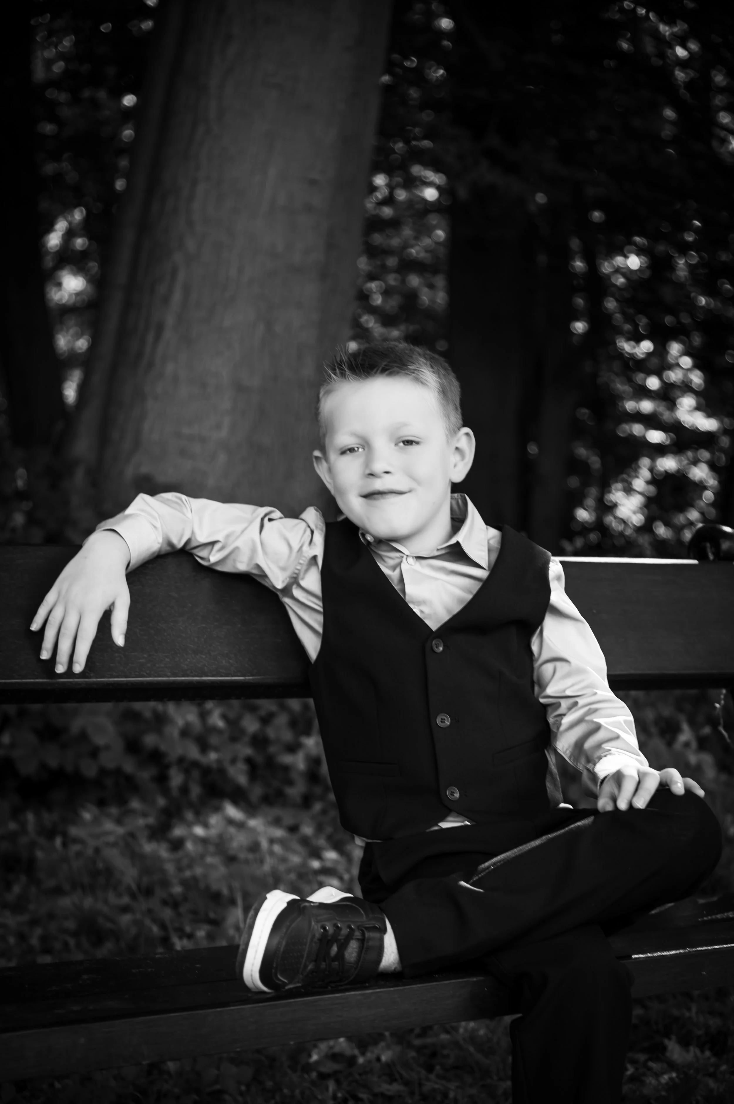 Zwarte en witte foto van een jonge jongen die op een bankje zit, met een grote boom en bomen op de achtergrond.