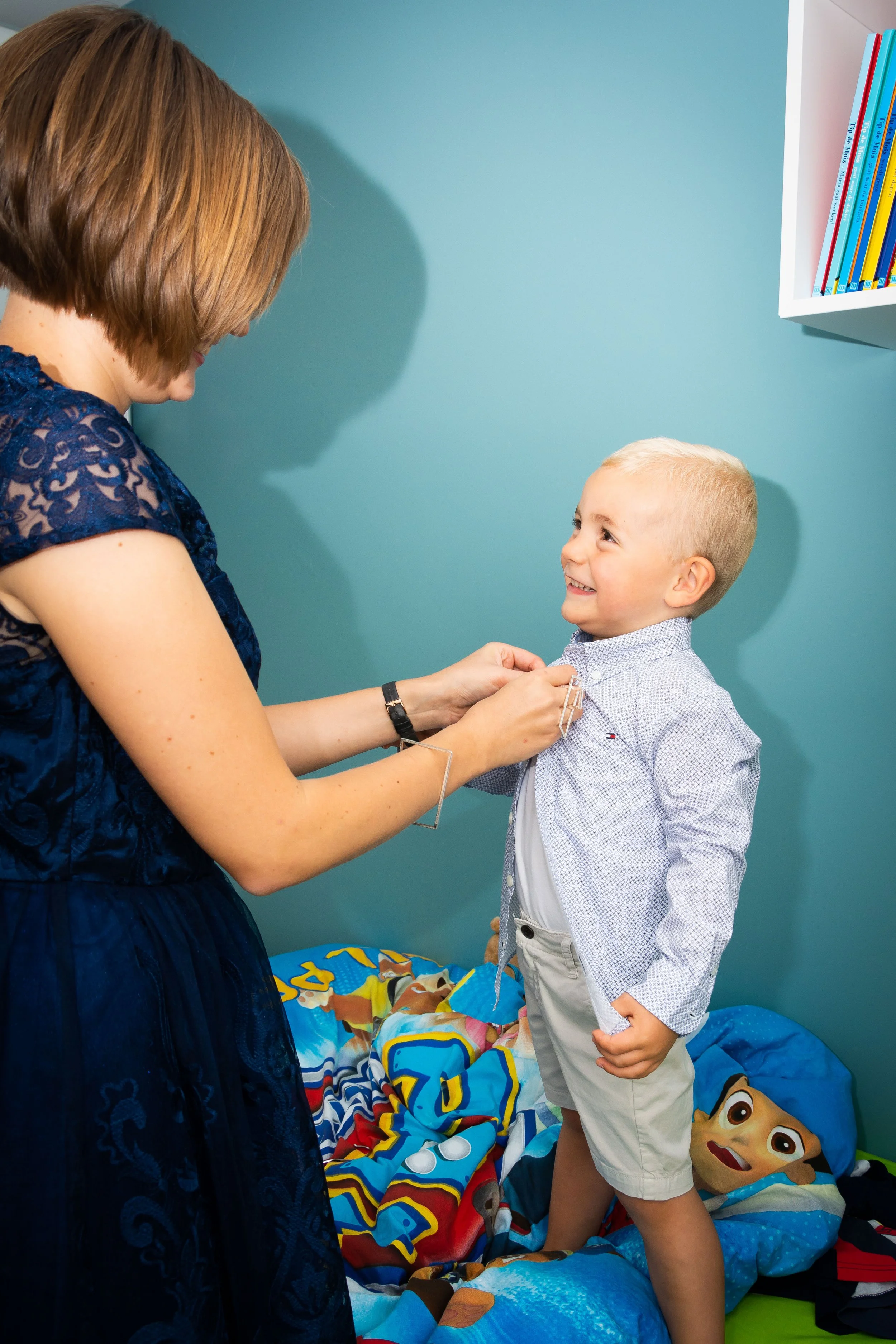 Een vrouw zet een jongen met blond haar en een breed lachend gezicht klaar voor een prik, terwijl hij op zijn bed staat met een Pyjama en een dekbed met pokemon thema.