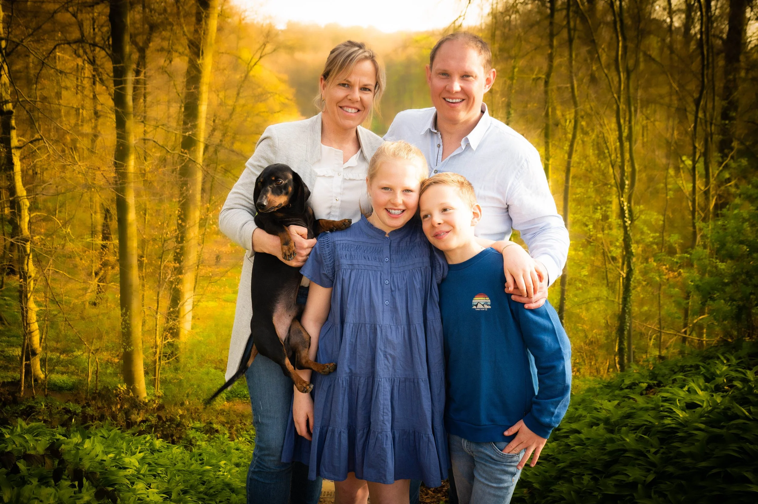 Gezinntje van vijf mensen met twee kinderen, een moeder, een vader en een puppy in een bos tijdens de herfst, allemaal lachend en knuffelend.