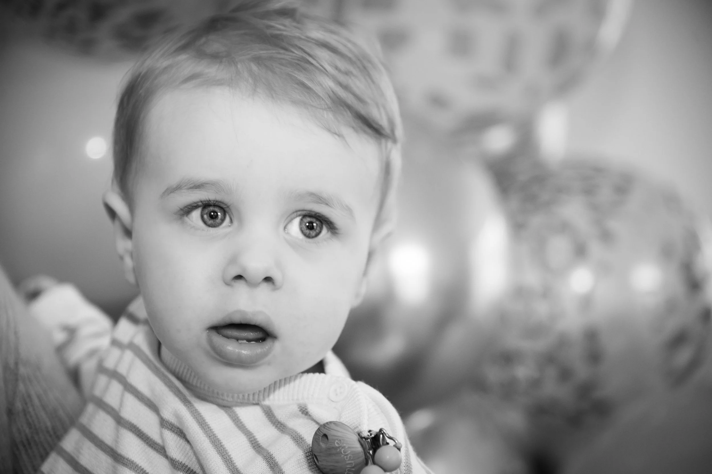 Zwart-wit foto van een jonge jongen met grote ogen die naar de camera kijkt, met een lichtgekleurde, gestreepte trui en een speen met een shakertje eraan.