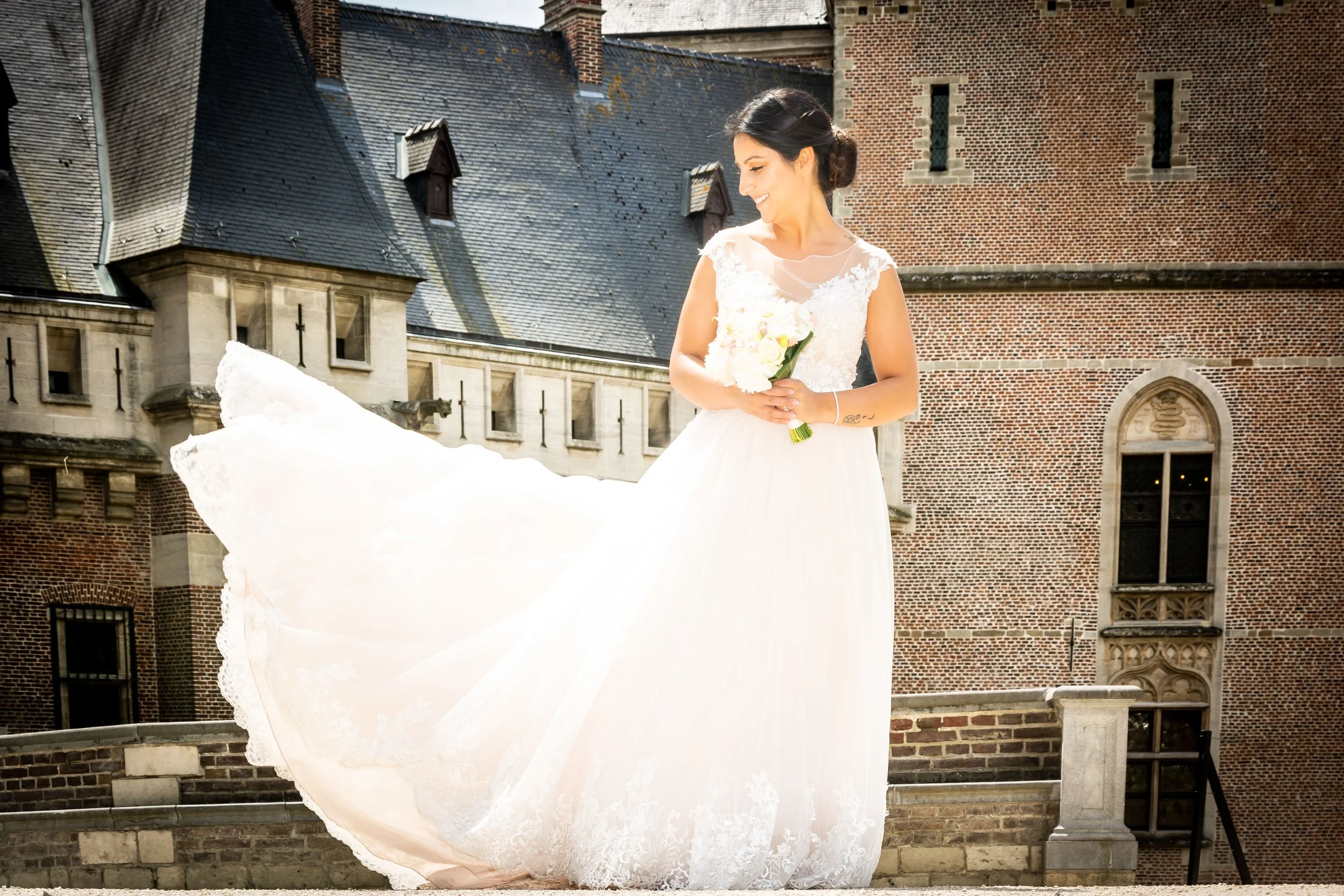 Vrouw in een witte trouwjurk met bloemen, staand buiten voor een kasteelachtige gebouw.