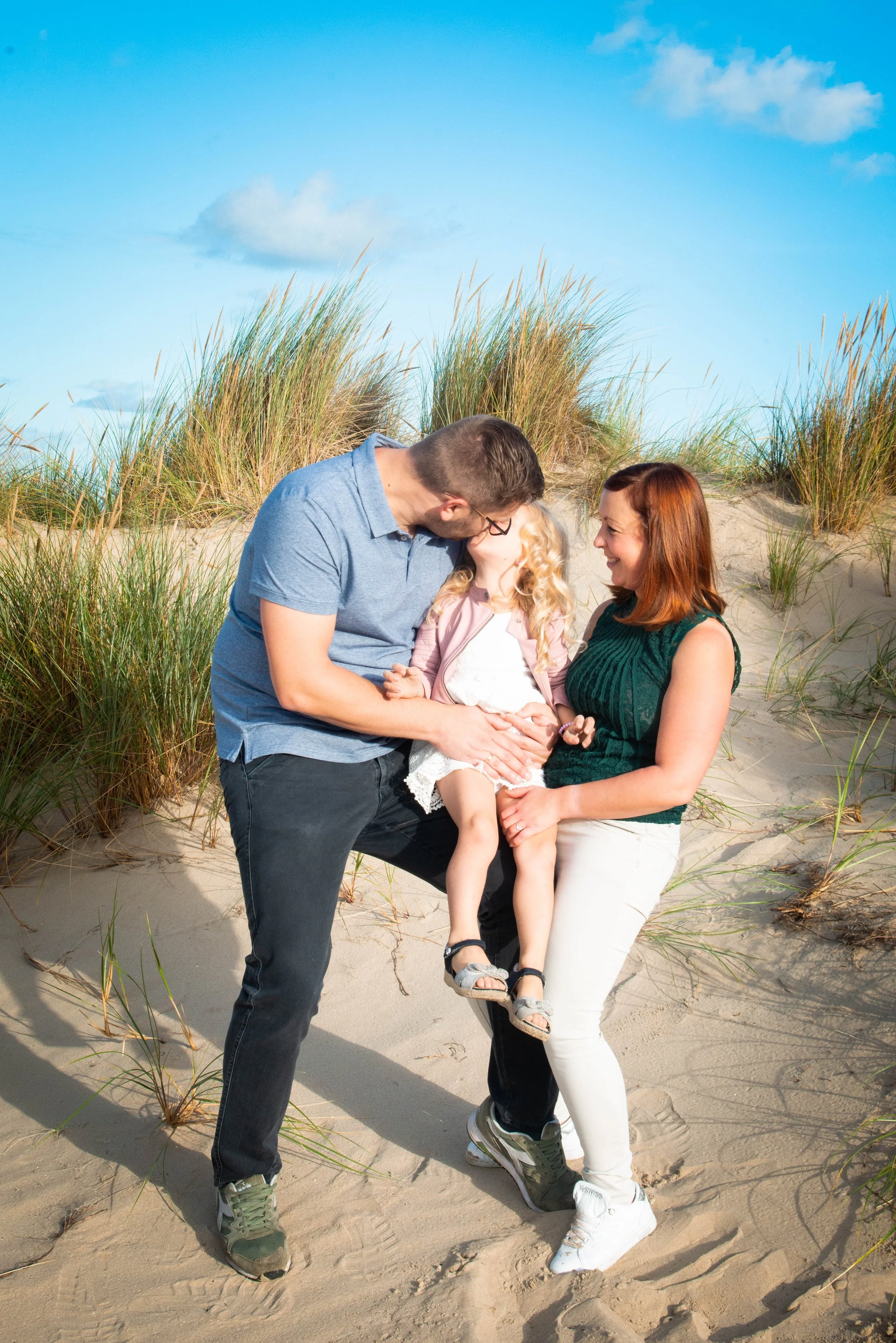 Een gezin op het strand met een man en vrouw die hun twee jonge dochters vasthouden, omgeven door duinen en gras, onder een blauwe hemel.