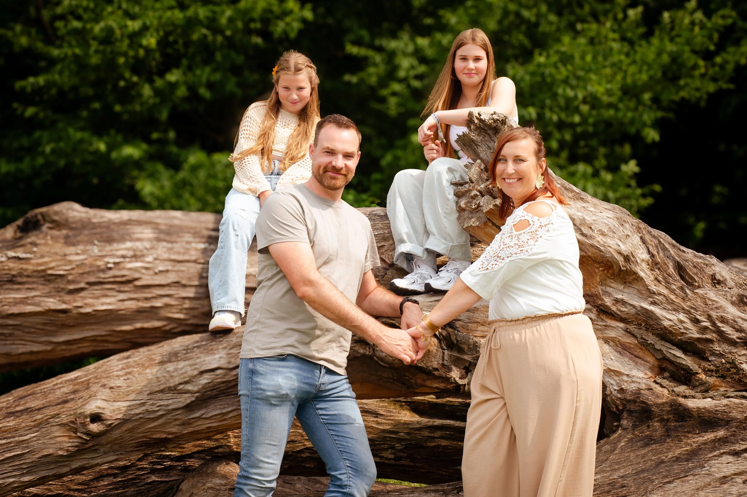 Een gezin bestaat uit twee volwassenen en twee kinderen die op een grote omgevallen boom zitten in een groene natuurlijke omgeving. De man en vrouw staan voor de boom en houden elkaars handen vast, terwijl de kinderen op de boom zitten.
