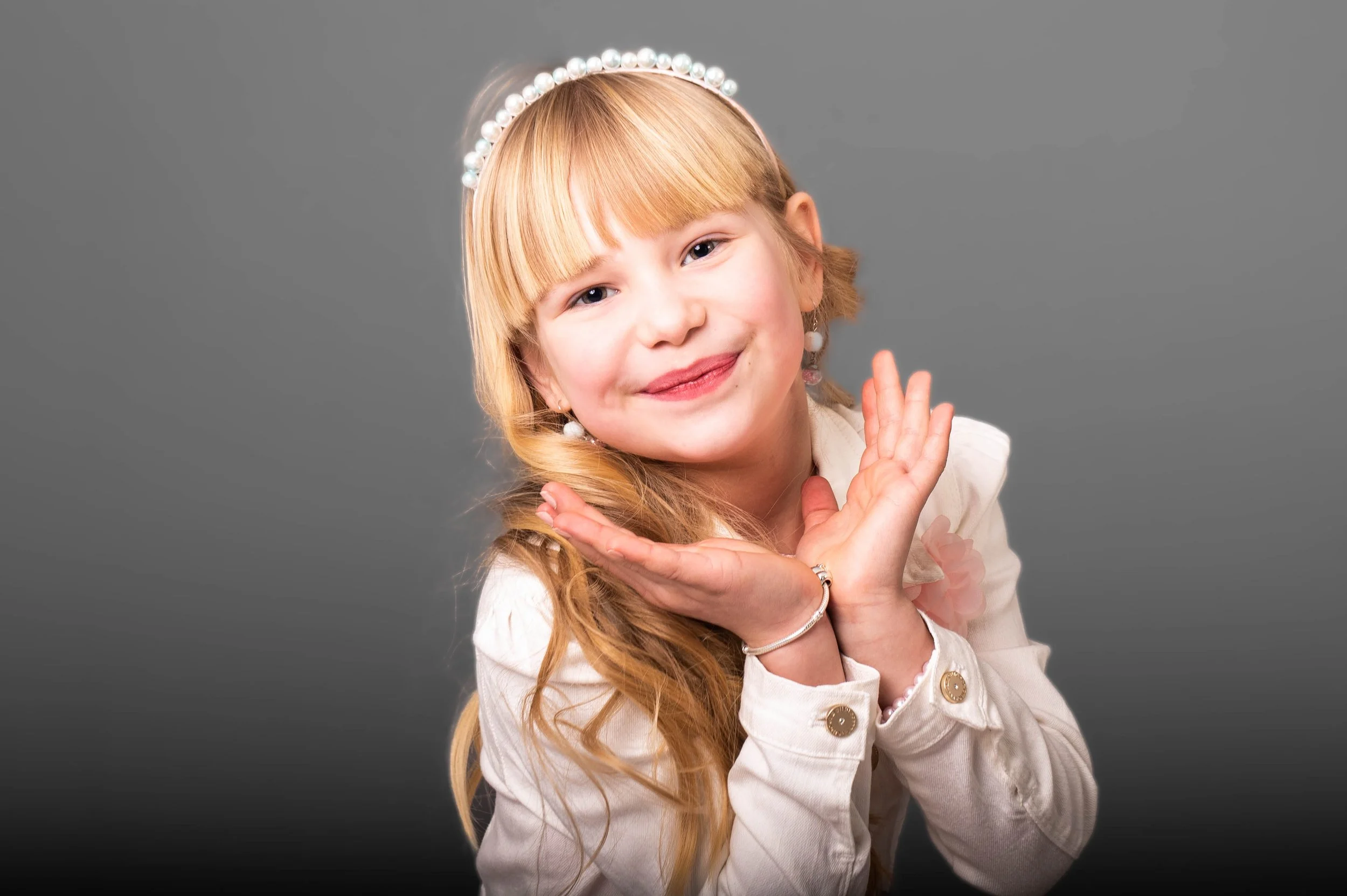 Een jonge meisje met lang, lichtbruin haar en vlechtjes, gekleed in een witte jas met gold-accenten, die lachend poseert met haar handen onder haar kin tegen een grijze achtergrond. Ze draagt een parelhoed en oorbellen.