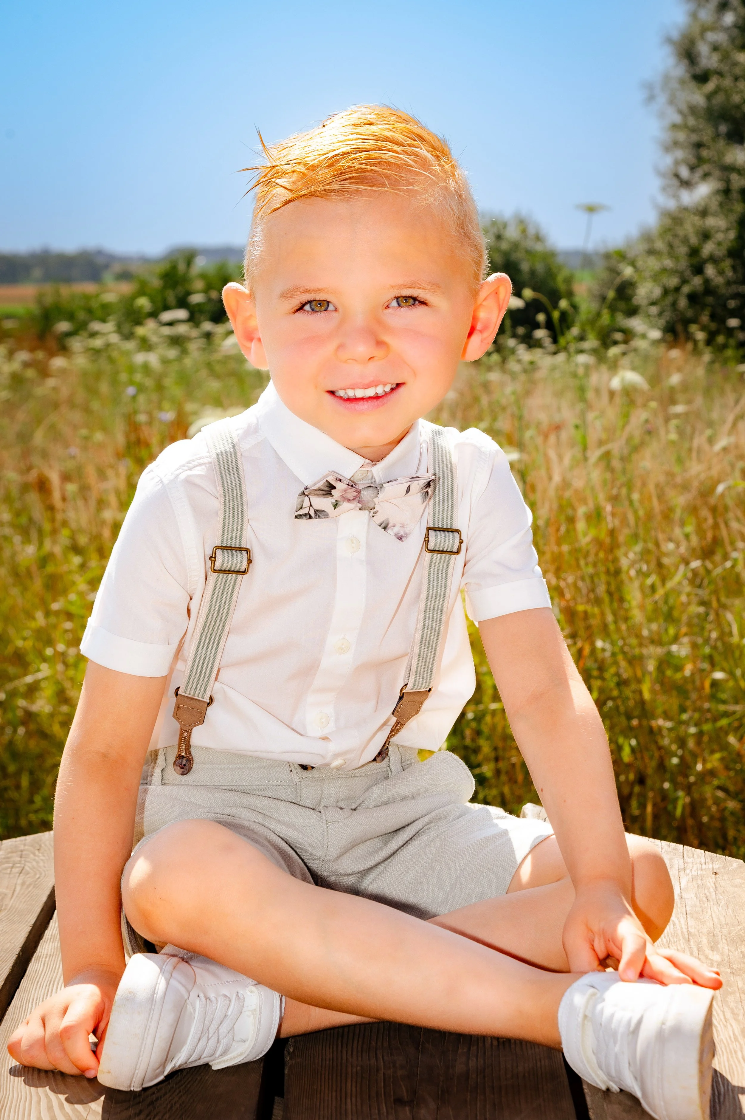 Een jonge jongen met blond haar, gekleed in een wit overhemd met een vlinderdas, beige shorts en witte sneakers, zittend op een houten platform in een veld met hoog gras en bloemen onder een blauwe lucht.