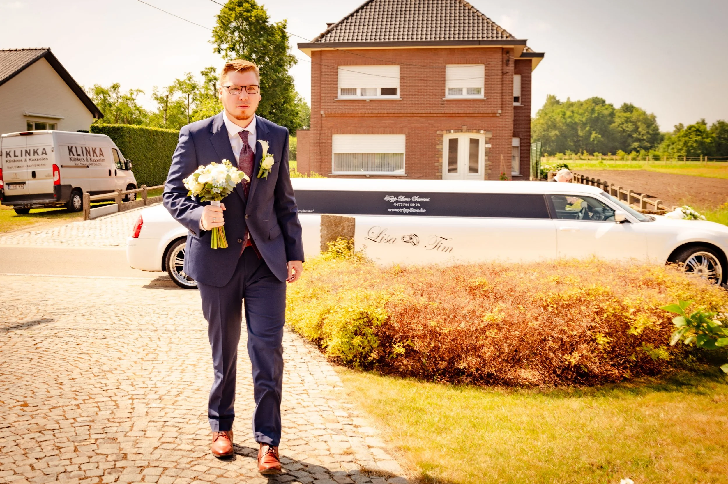 Man in een donkerblauw pak met stropdas en bruine schoenen, wandelend met een boeket witte en groene bloemen voor een trouwauto. Achtergrond een huis, een gebouw met bedrijfswagens, en een burgerlijke limousinewagen met teksten 'Lisa & Tim'.