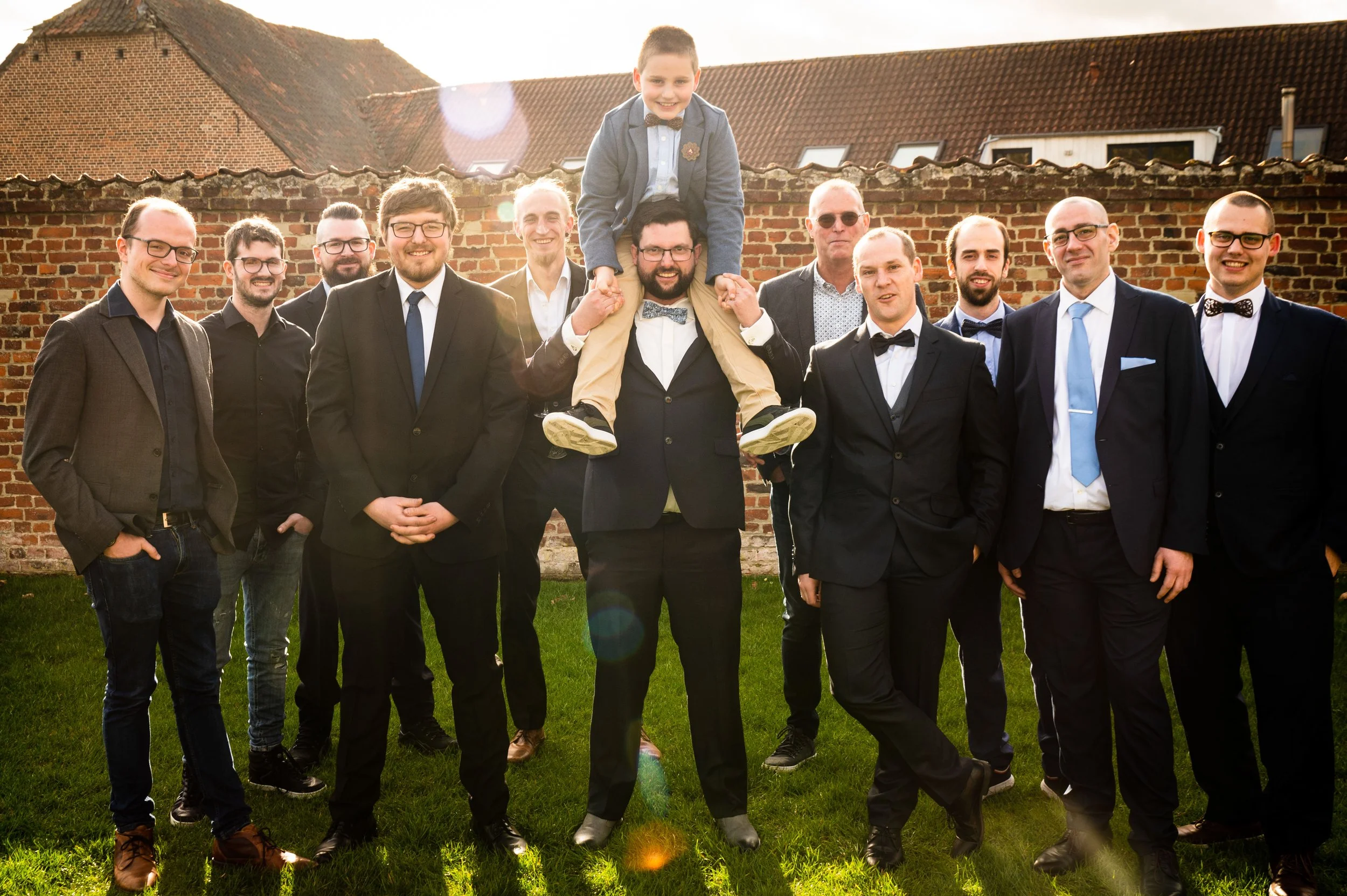 Vrolijk gezelschap in formele kleding, waaronder enkele in stropdassen en bretels, poseert buiten voor een bakstenen muur met een zonnige achtergrond.