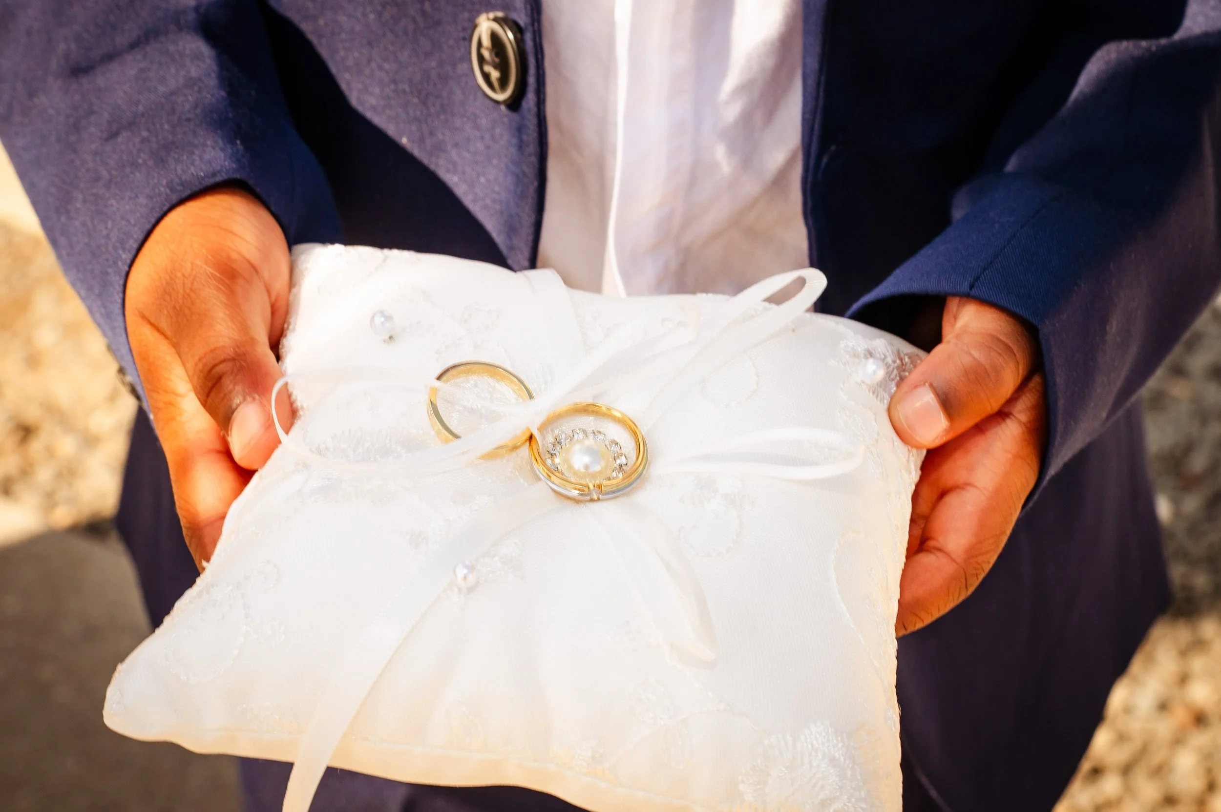Persoon houdt een witte satijnen ringkussen met twee gouden huwelijksringen en een parelversiering erop.