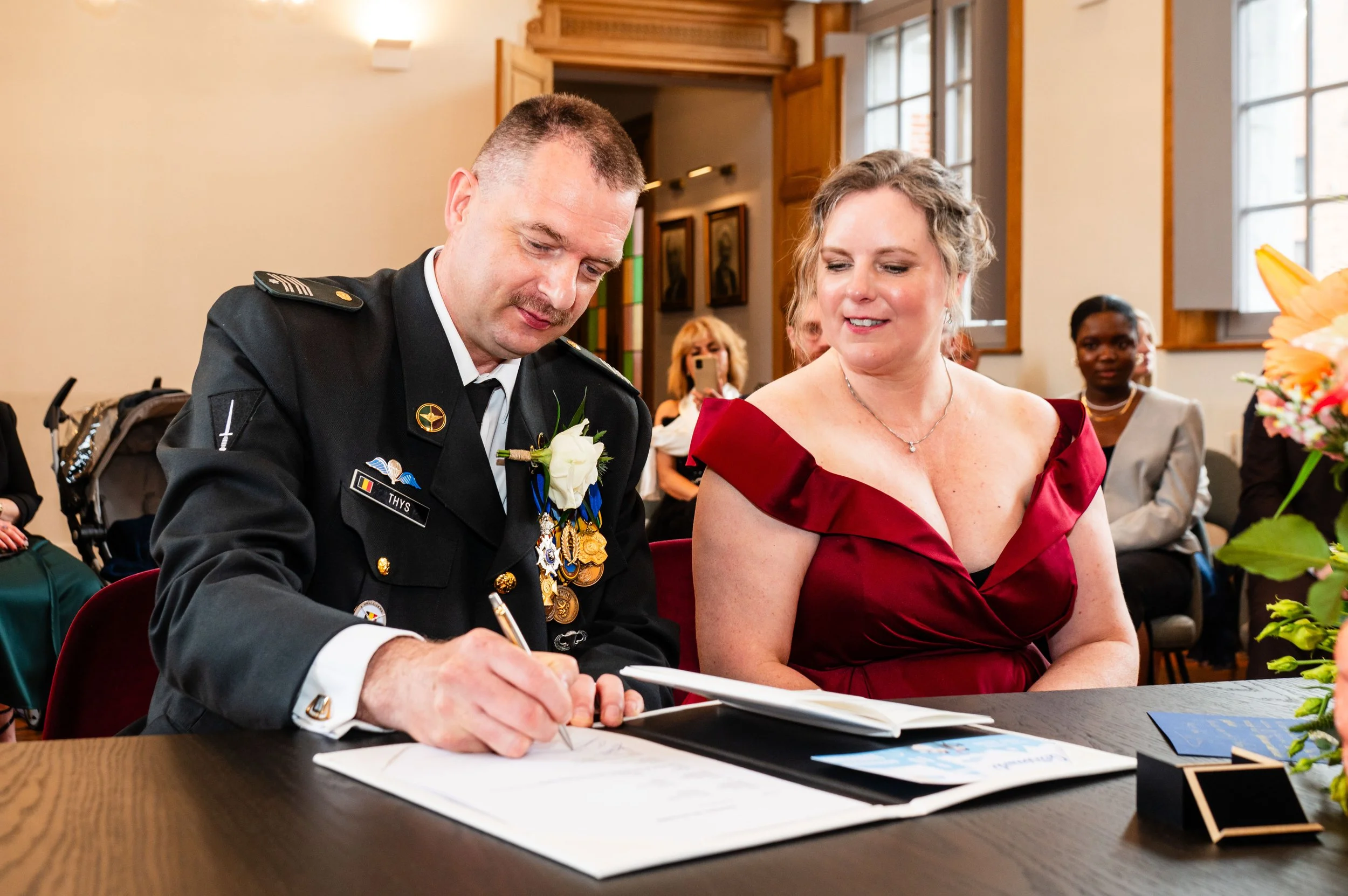 Een man in een militaire uniform ondertekent een document terwijl een vrouw in een rode jurk naast hem kijkt tijdens een ceremonie.