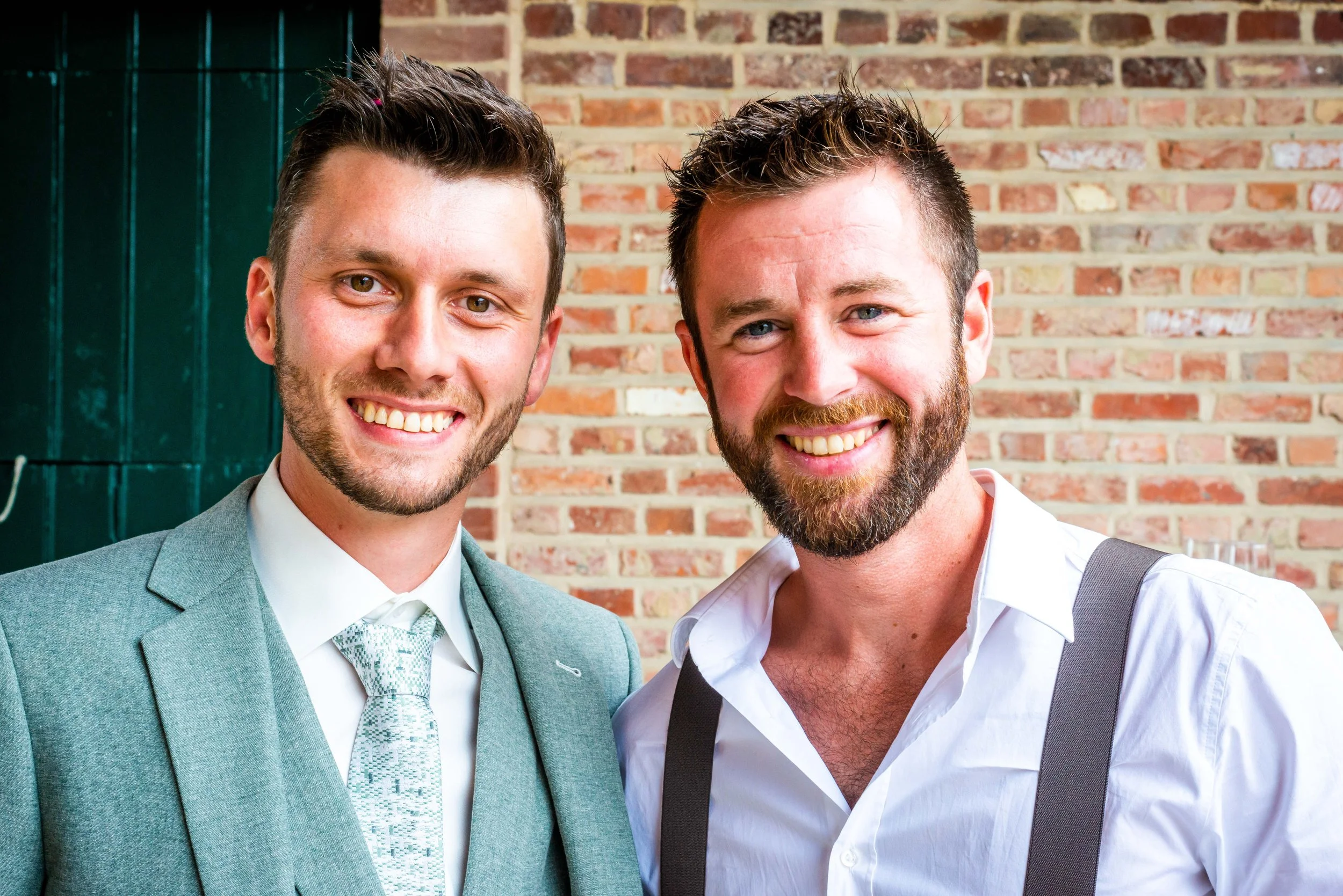 Twee mannen met glimlach staan voor een bakstenen muur, een in pak en een in een witte shirt met een das en een suède schouderriem.