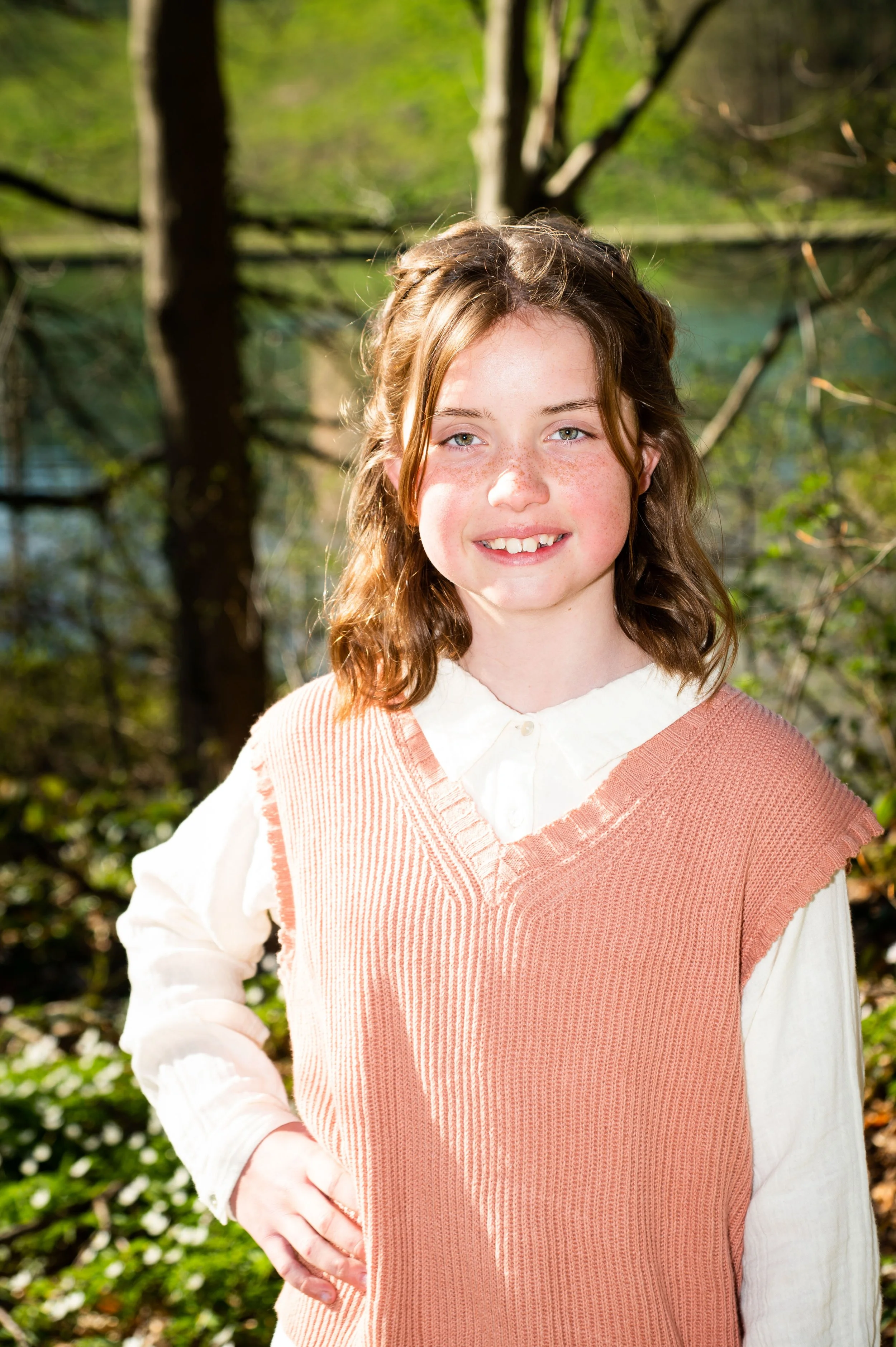 Een lachende jonge vrouw met bruin haar in een bos, gekleed in een wit shirt en een zalmroze gebreide vest.