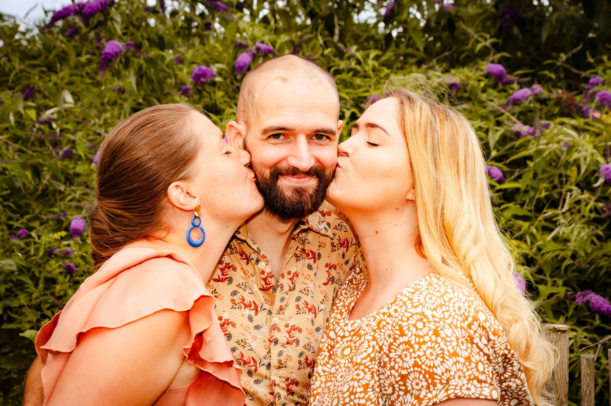 Twee vrouwen kussen een man op de wangen terwijl ze in een tuin met paarse bloemen staan.