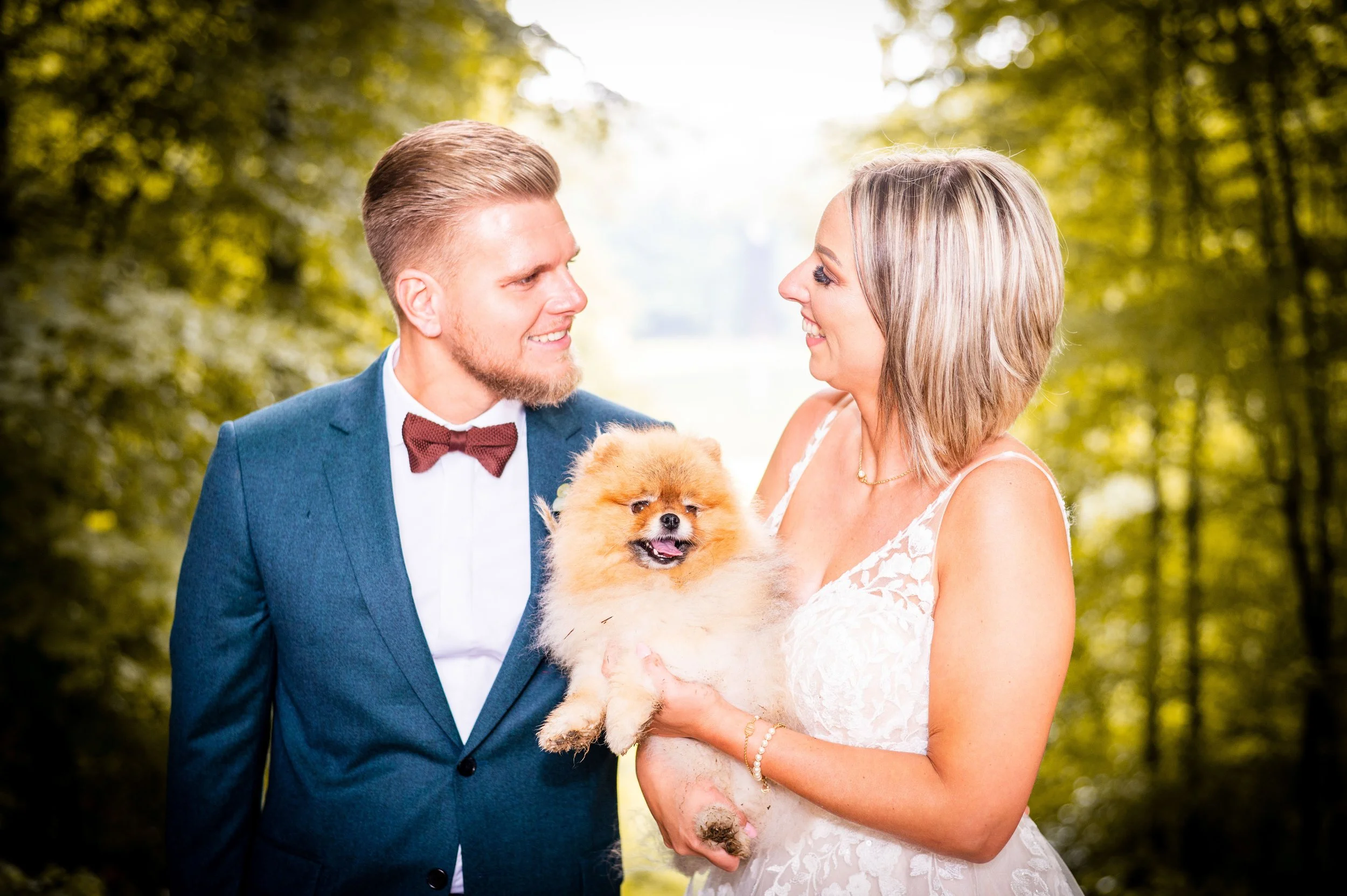 Een bruid en bruidegom in een bos, samen met een klein, fluffy hondenras, mogelijk een Pomeranian, terwijl ze naar elkaar glimlachen.