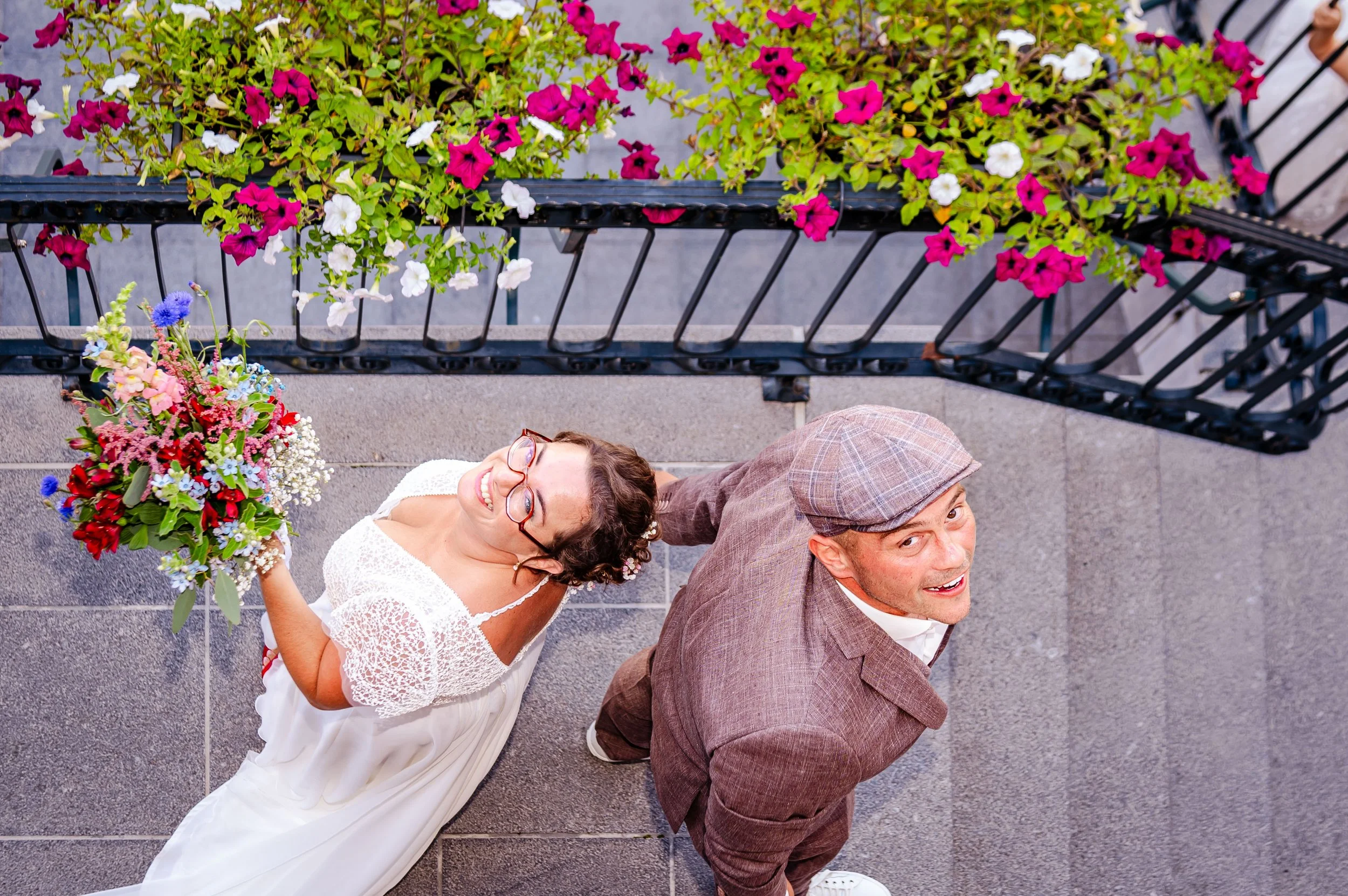 een vrouw met een witte jurk en een man in een bruin geruit pak staan op tegels, de vrouw houdt een grote bloemenbundel vast, en ze kijken naar boven en lachen op een ronde foto vanaf een hoog punt genomen met bloemen en hekwerk eromheen.