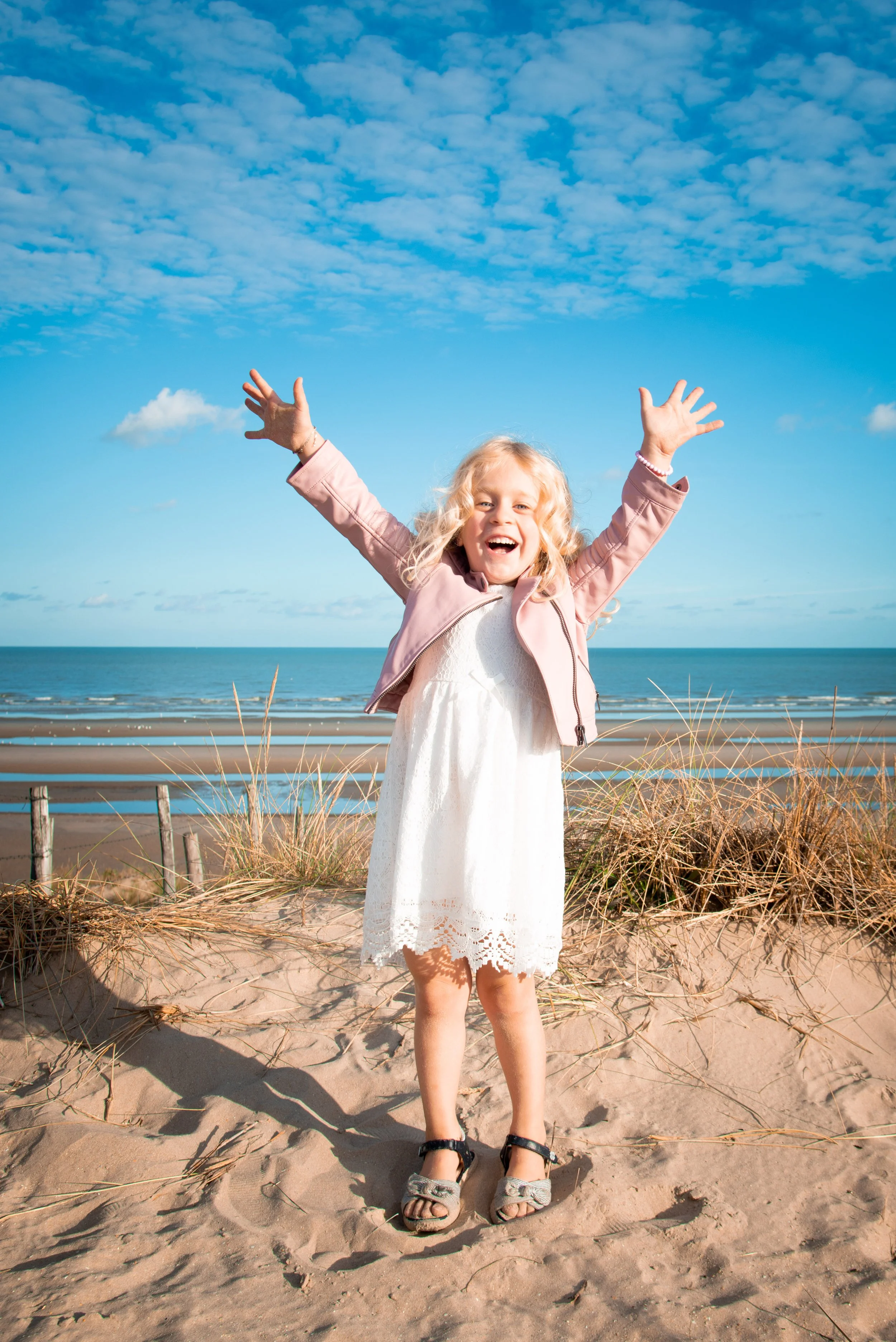 Lachende jonge meisje met roze jas en witte jurk op zandstrand, met handen omhoog, blauwe lucht en oceaan op de achtergrond.