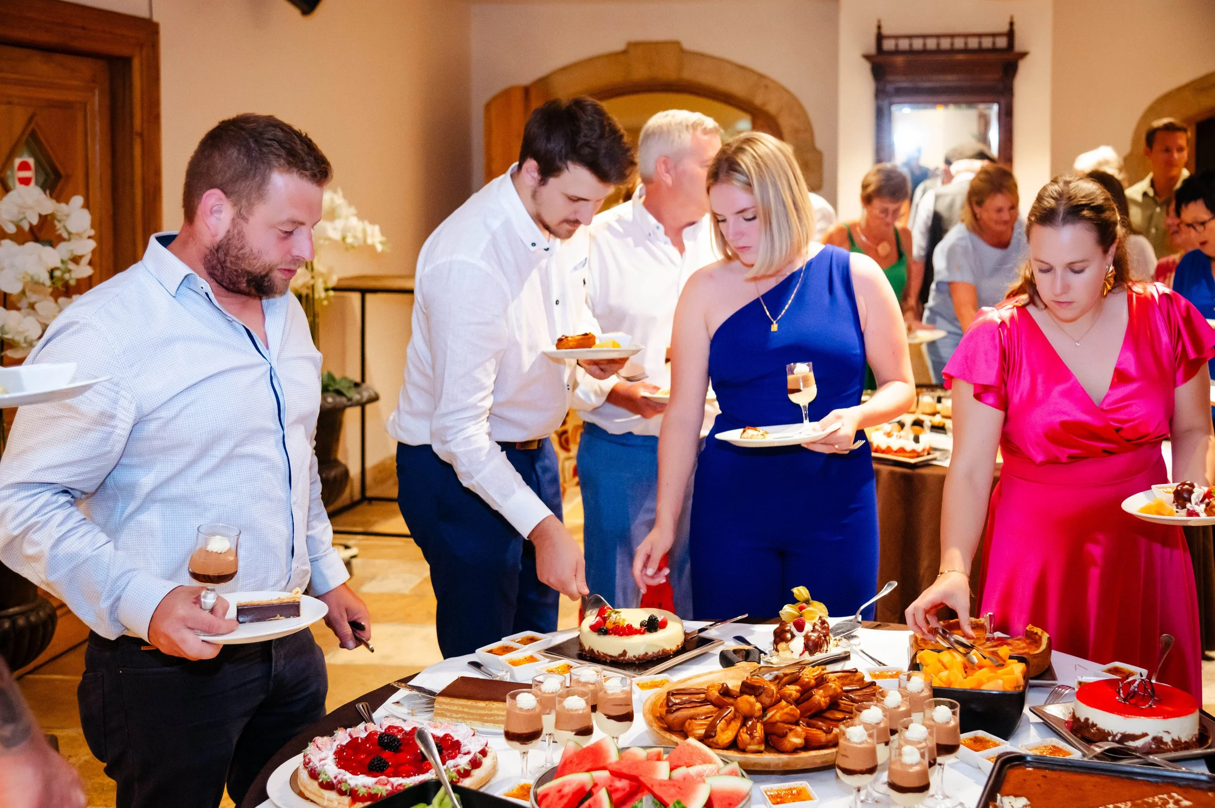 Mensen op een feestbuffet met diverse desserts, zoals taart, fruit, en kleine chocolade desserts, in een gezellige, beige ingerichte ruimte.
