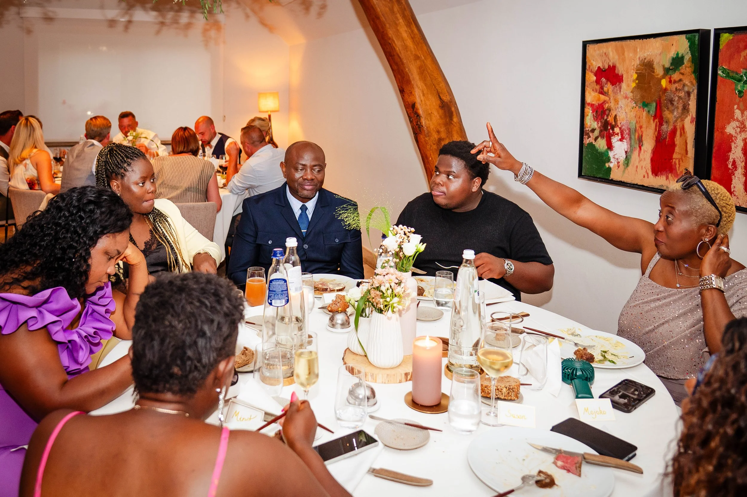 Mensen zitten rond een eettafel tijdens een diner, met één persoon die een sprekersrol heeft en een glimlach op het gezicht. Op tafel staan bloemen, drankjes, en borden met eten.