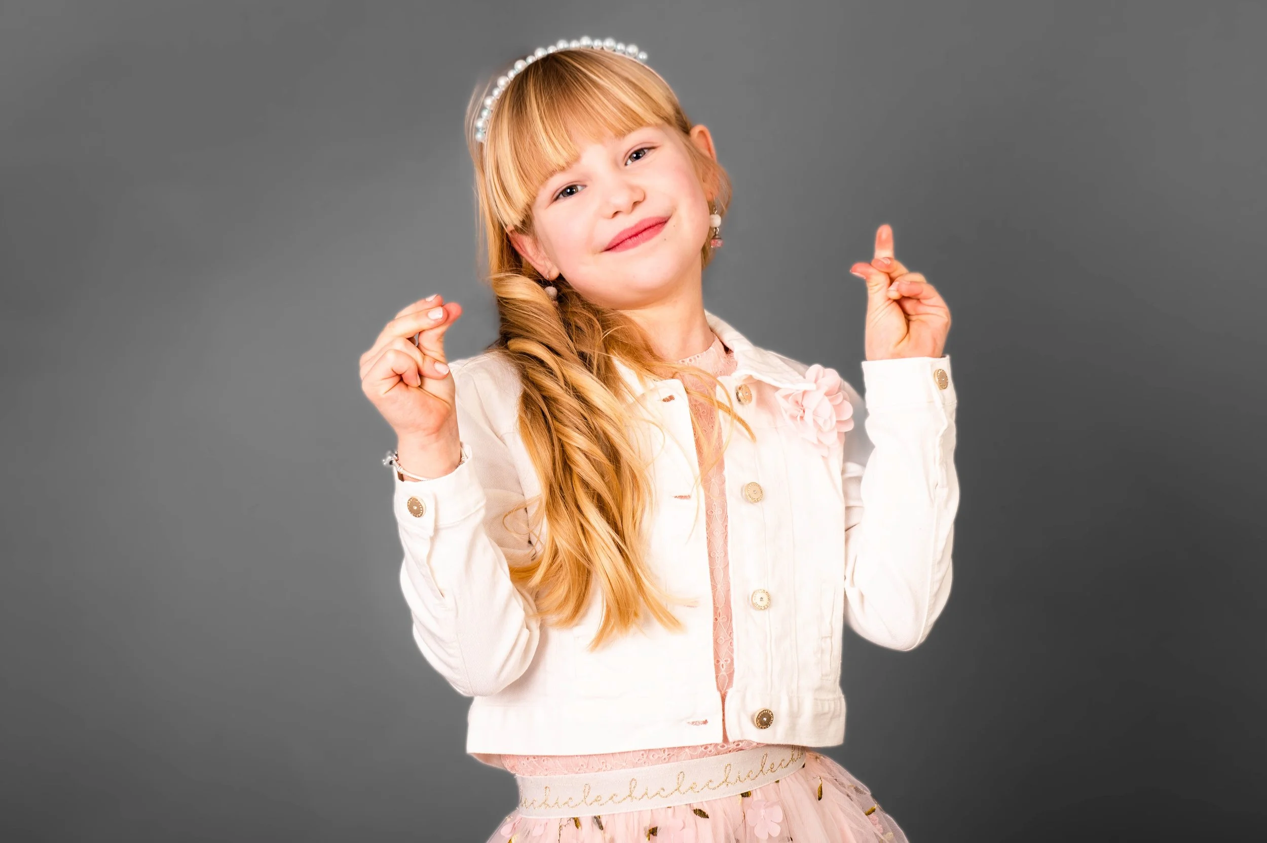 Lachende jonge meisje met lange blonde haar, gekleed in een witte jas en een roze jurk, poseert tegen een grijze achtergrond.
