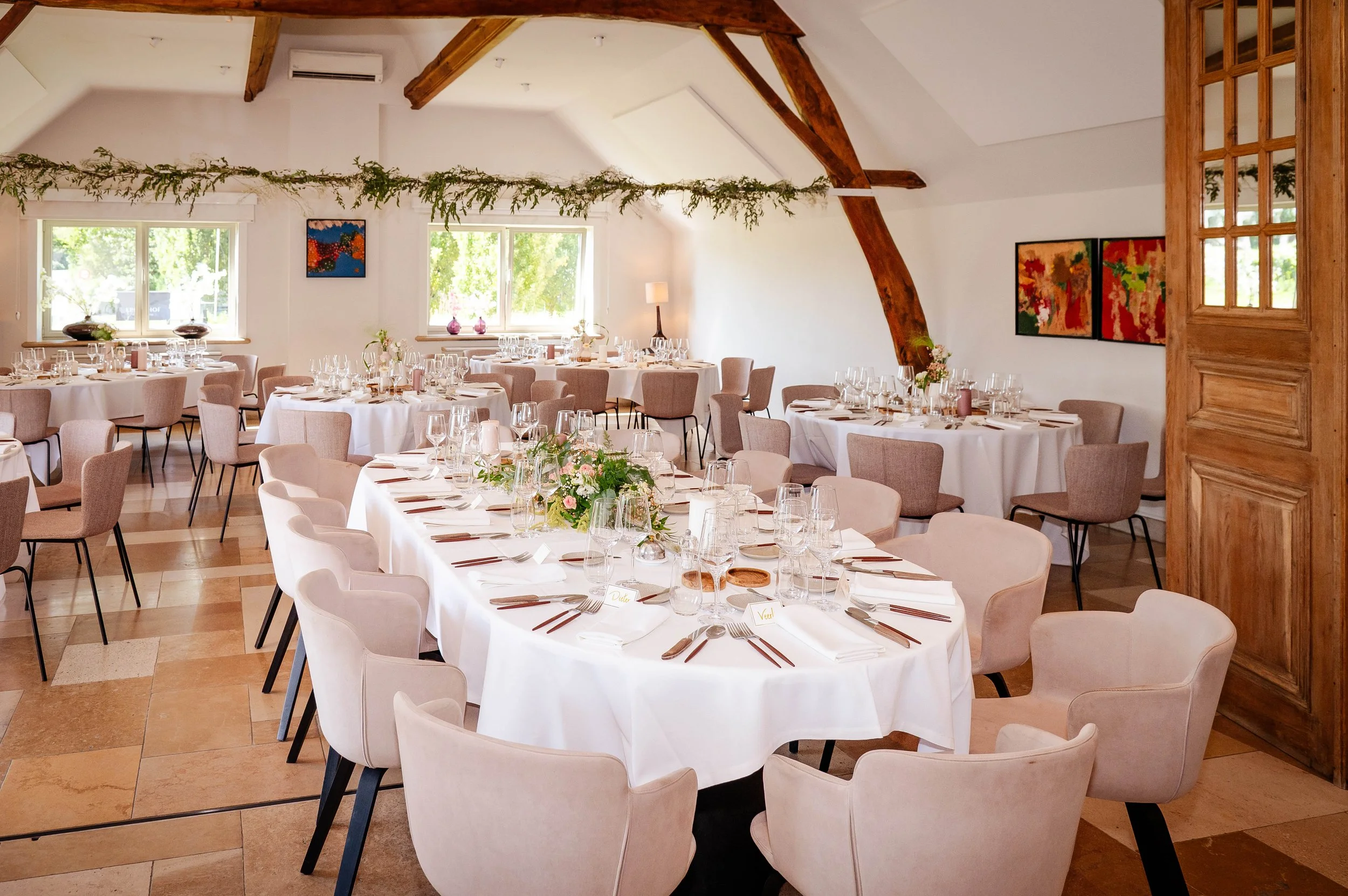 Tafelopstelling voor een formeel diner met witte tafelkleden, glaswerk, bestek en bloemstukken in een feestzaal met grote ramen, houten balken en kleurrijke kunstwerken aan de muur.