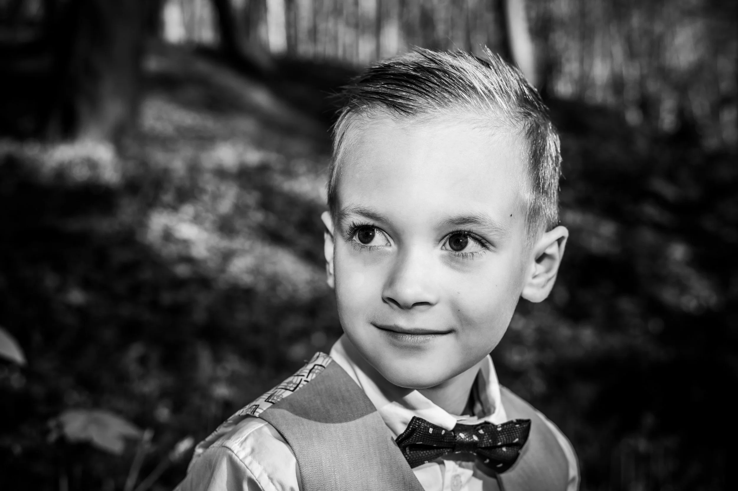 Zwarte en witte foto van een jonge jongen met een gestreken kapsel, gekleed in een overhemd en een vlinderdas, in een bos met bomen op achtergrond.