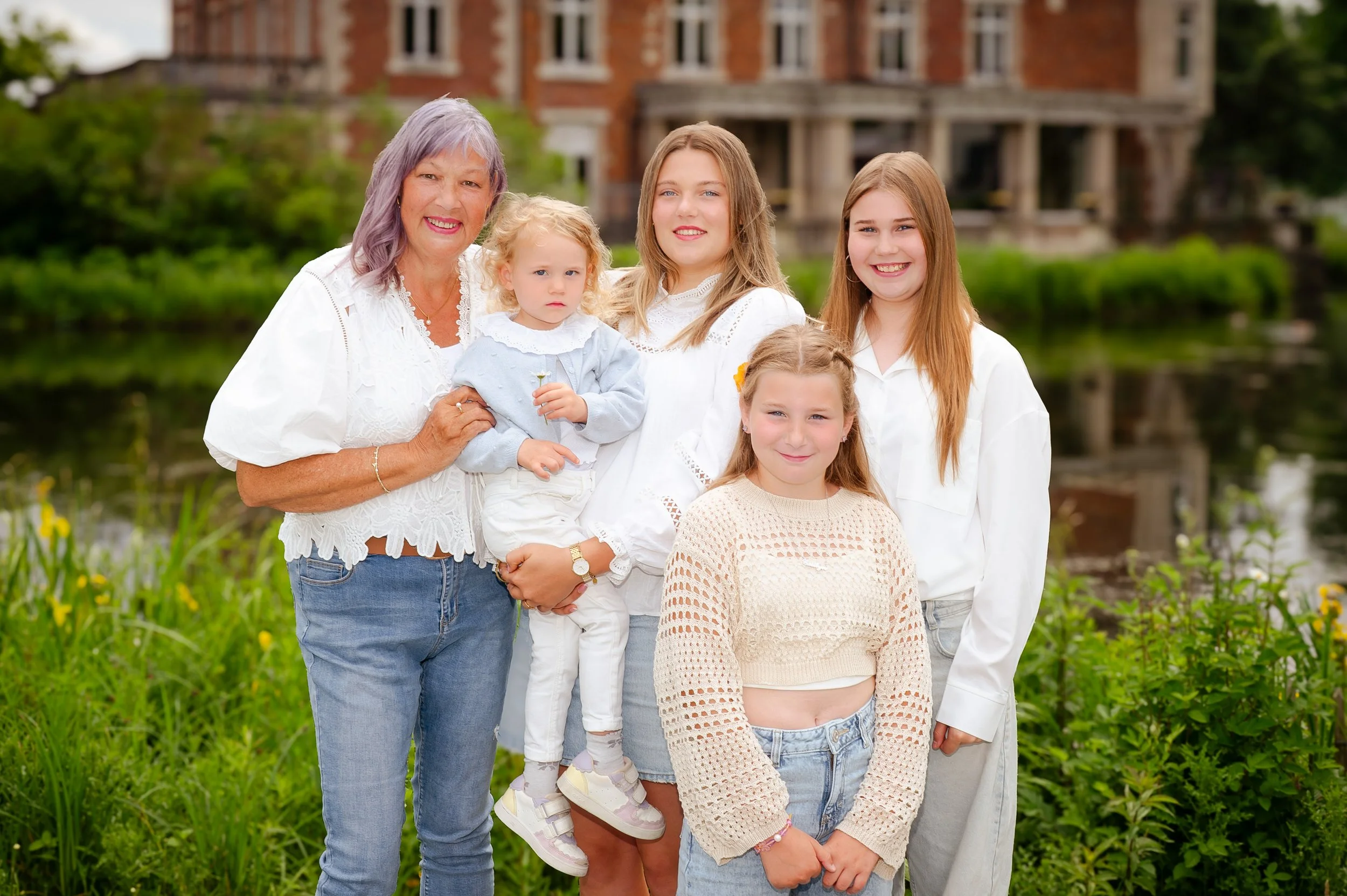 Vrouw met vier meisjes en een jonge meisje, buiten bij een rivier met een groot huis op de achtergrond, allemaal gekleed in witte of lichte kleding, lachend en poserend voor de camera.