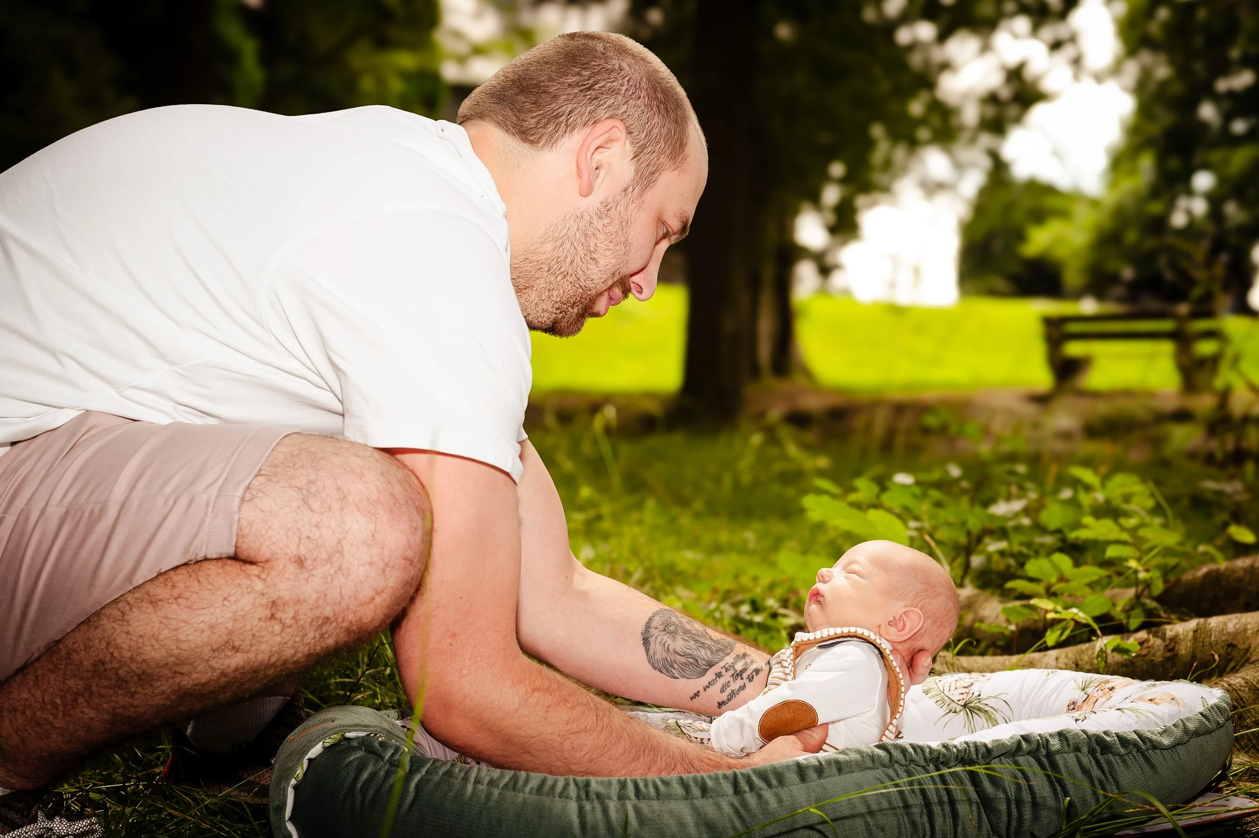 Een man knielt op de grond die in een park ligt, terwijl hij naar een slapende baby in een draagmandje kijkt.