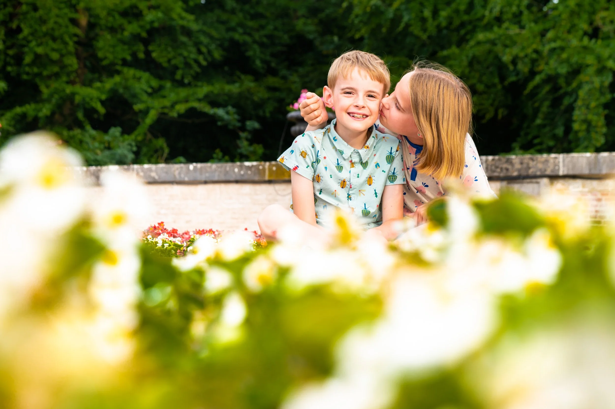 Een jongen en een meisje zitten op de grond tussen bloemen, de jongen wordt gekust door het meisje terwijl ze vrolijk lachen, met een groene achtergrond.